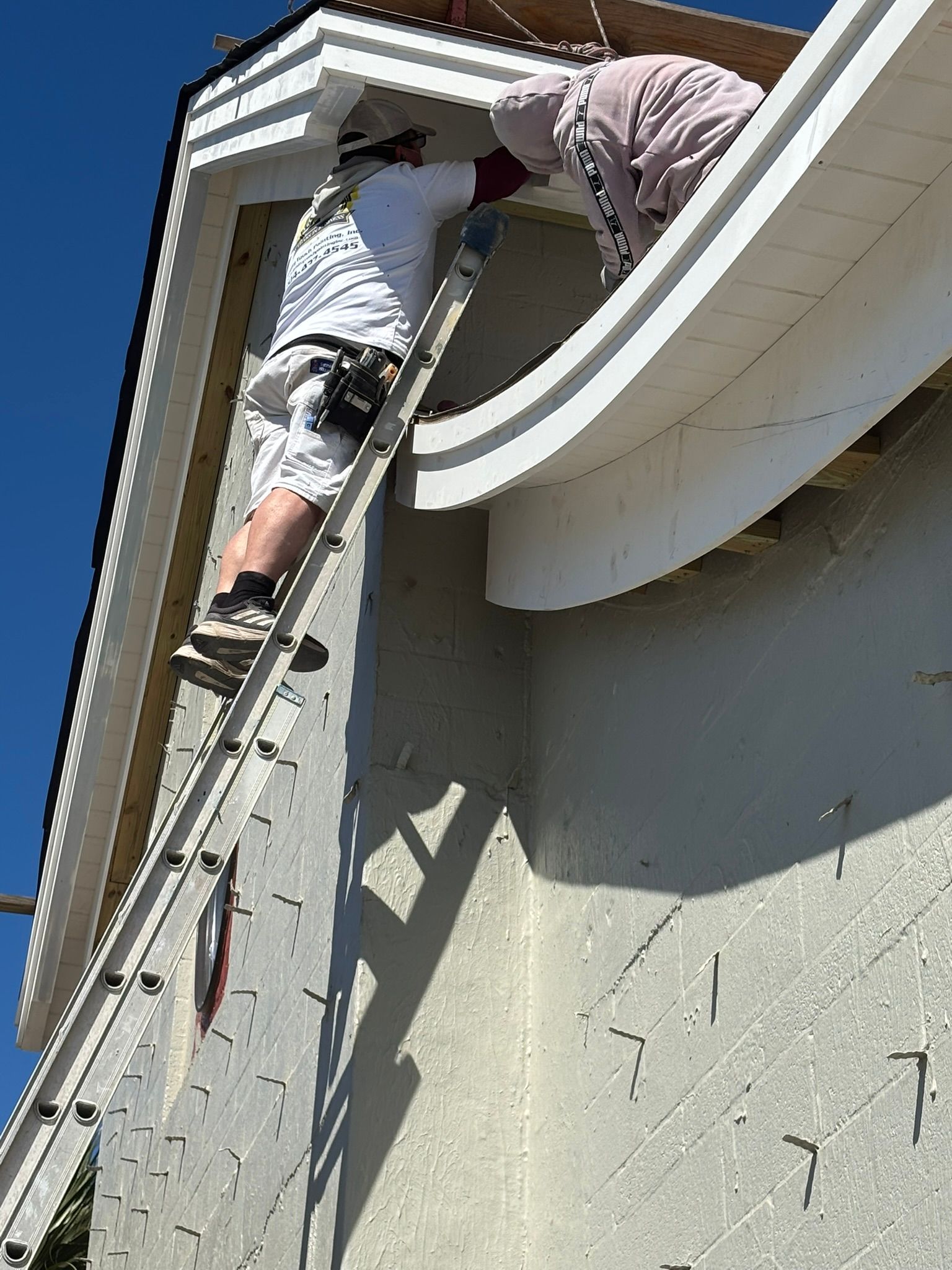 Two people painting a house exterior: one on a ladder, the other on the roof. Sunny day, light-colored building.