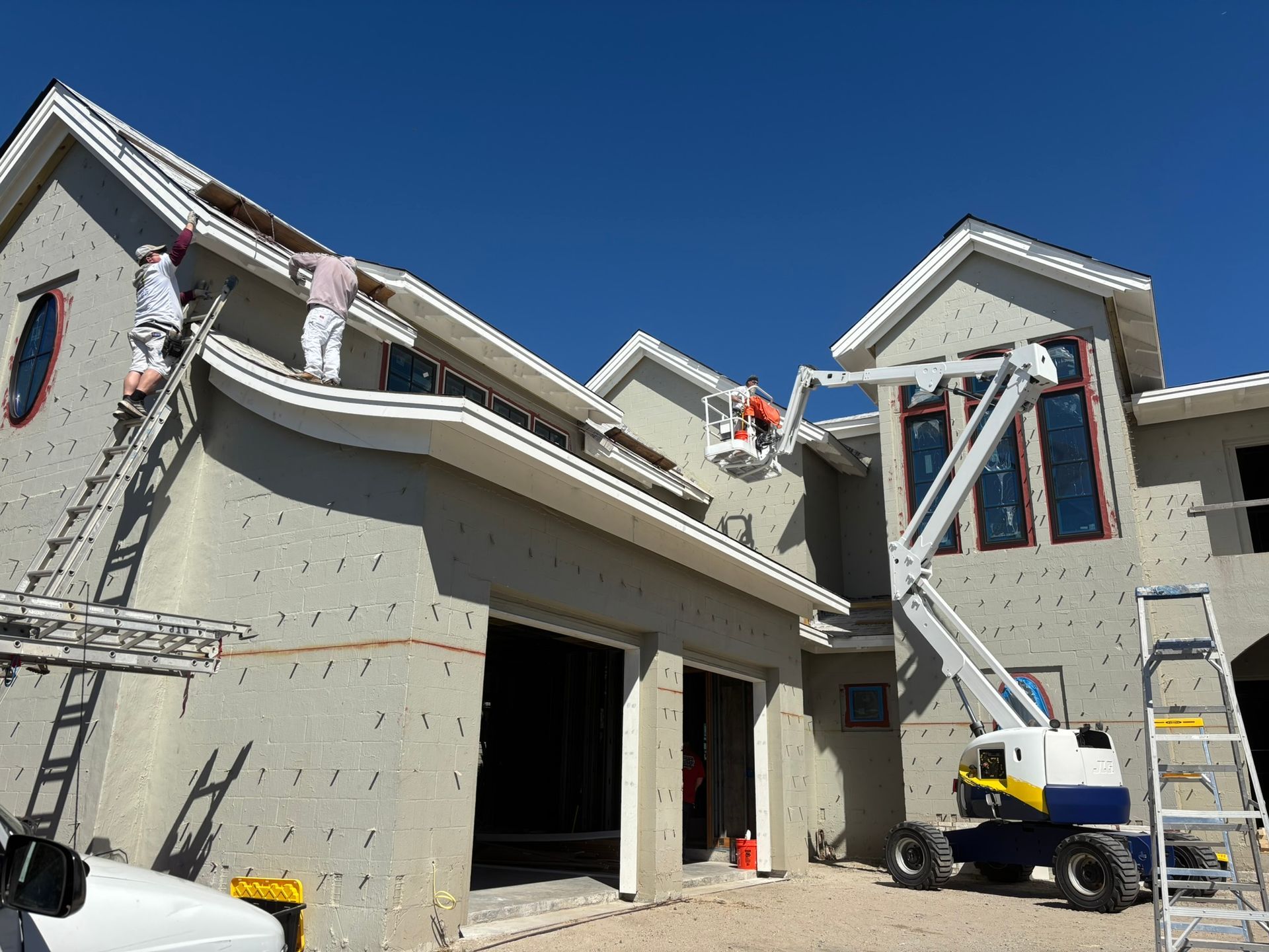 Workers painting the exterior of a large house, using ladders and a lift against a blue sky.