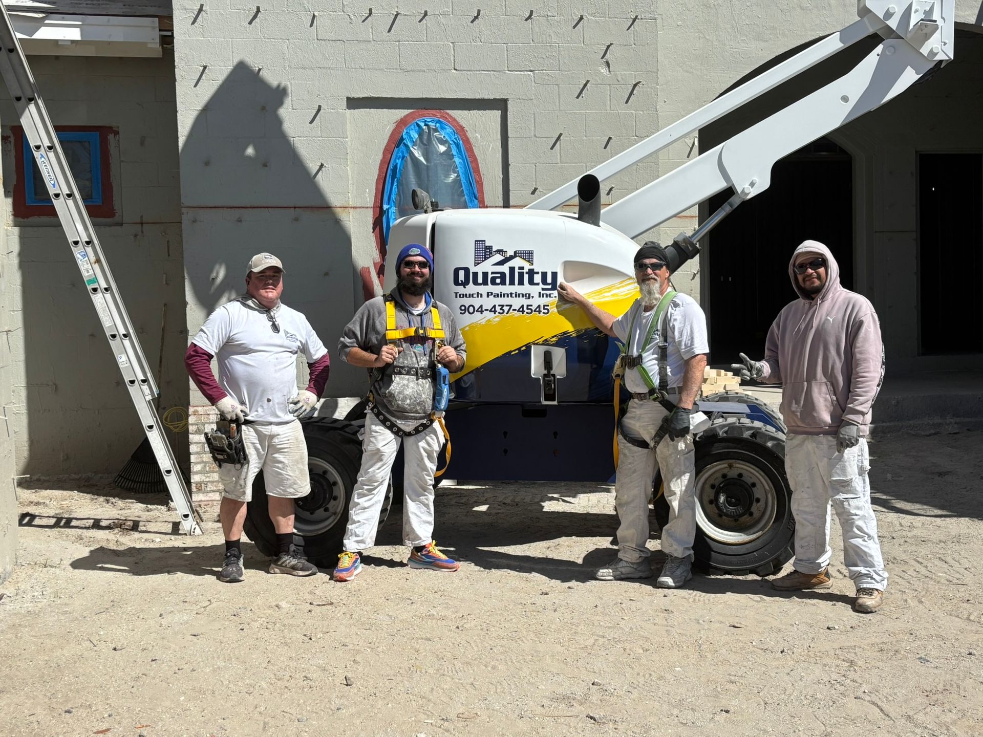 Four construction workers in front of a boom lift on a building site. They wear harnesses and work clothes.