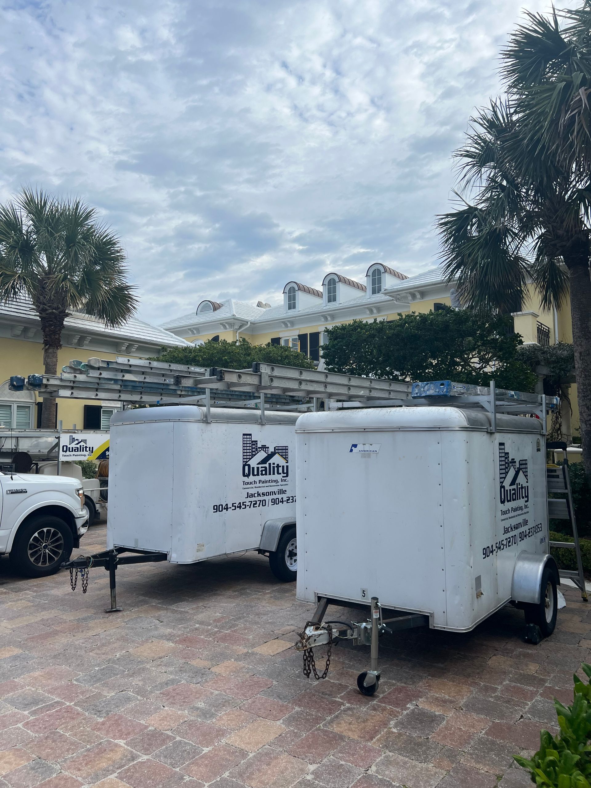 Two white utility trailers with ladders parked on brick near a building and trees under a cloudy sky.