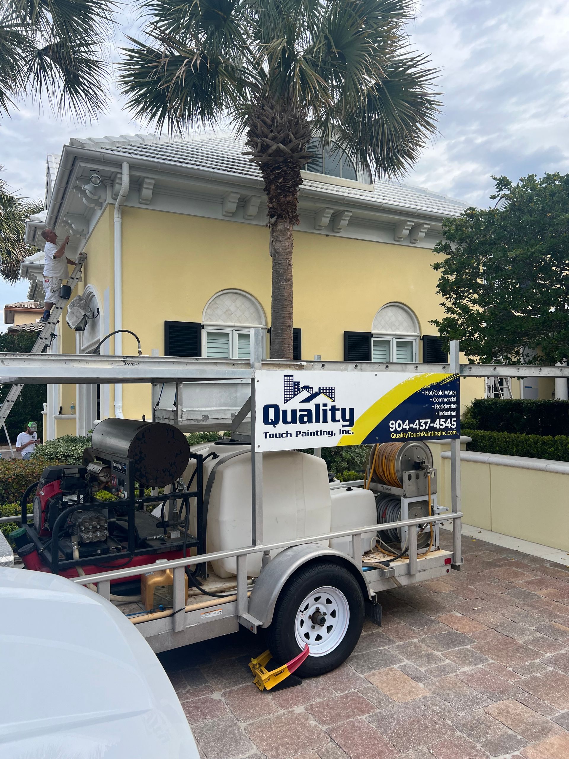 Painting crew working on a yellow house from a trailer-mounted pressure washing system near a palm tree.