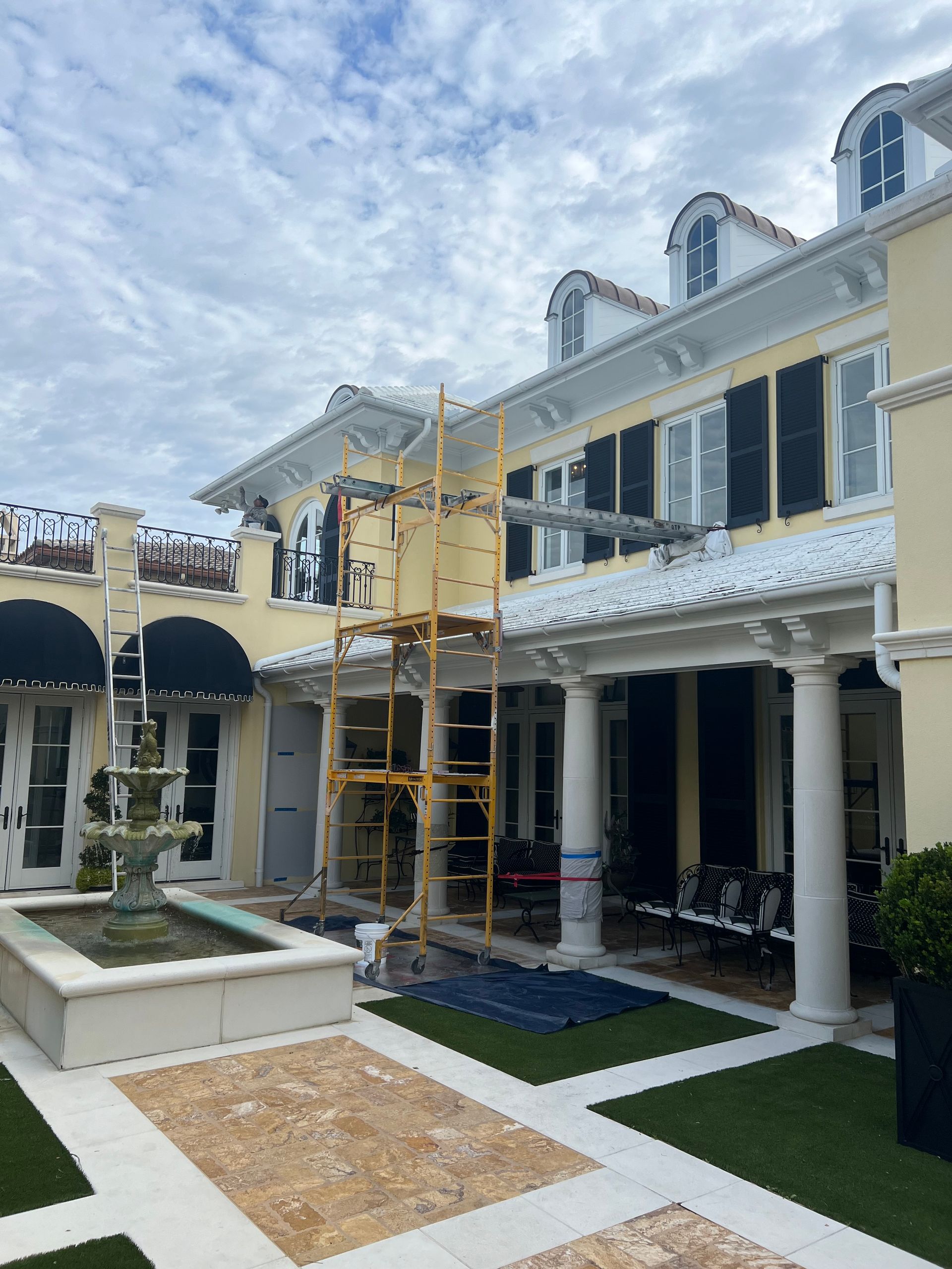 Exterior of a yellow mansion with scaffolding, undergoing renovations. A fountain sits in the foreground, with cloudy sky above.