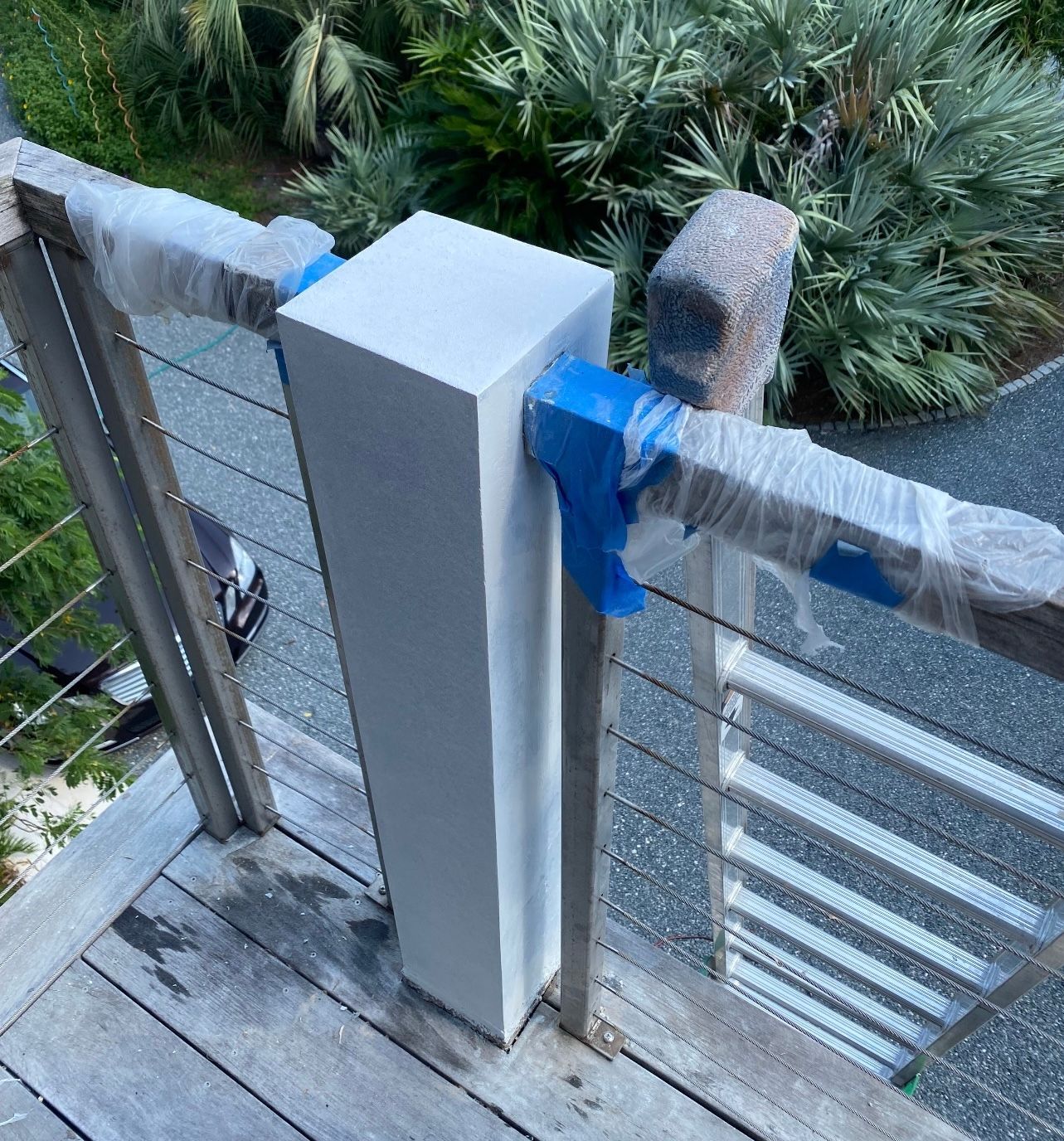 White pillar with wooden railing, secured with tape, on a deck. Stainless steel cables and green foliage in the background.