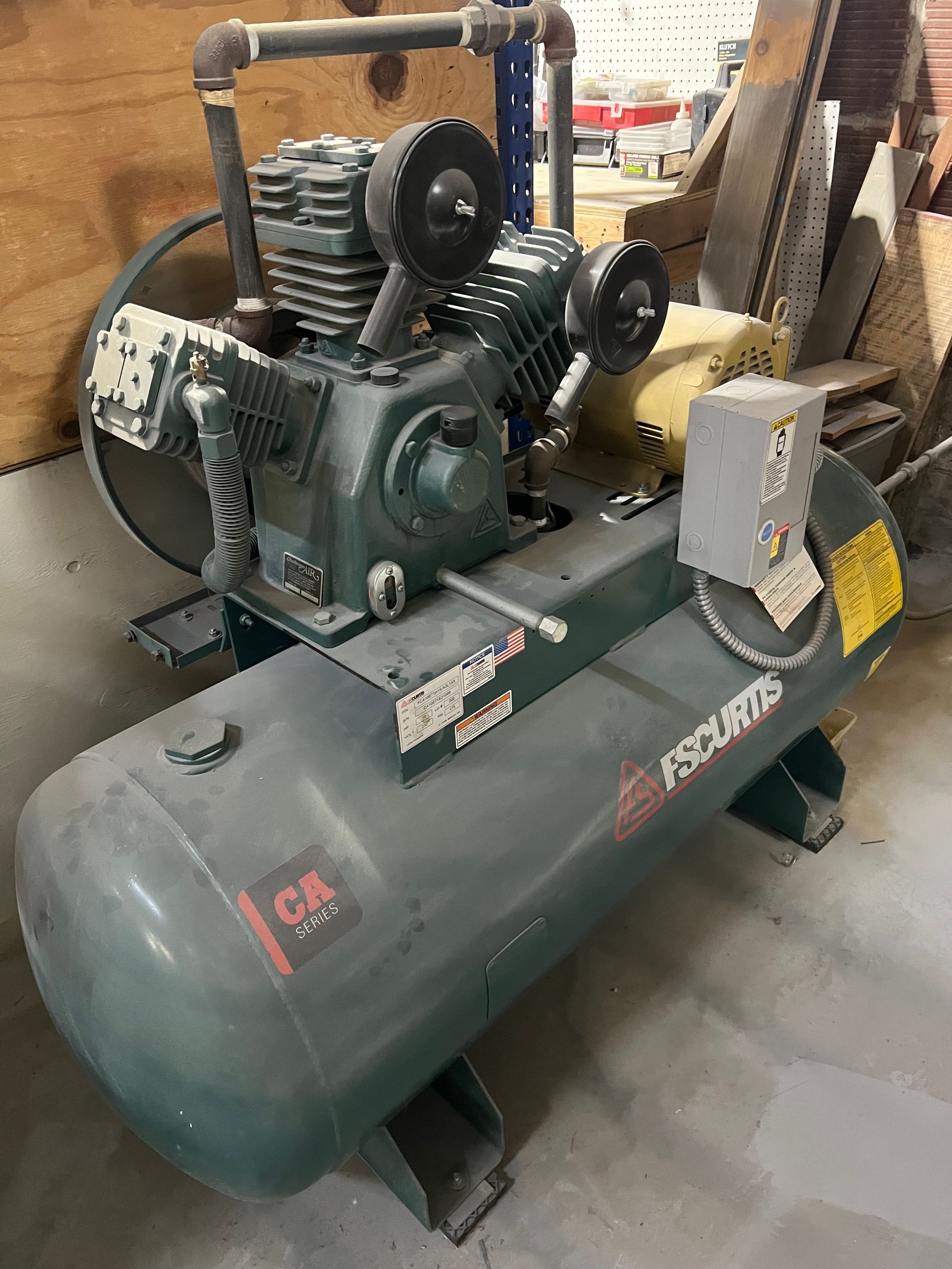 A green industrial air compressor with a cylindrical tank, metal piping, and gauges in a workshop.