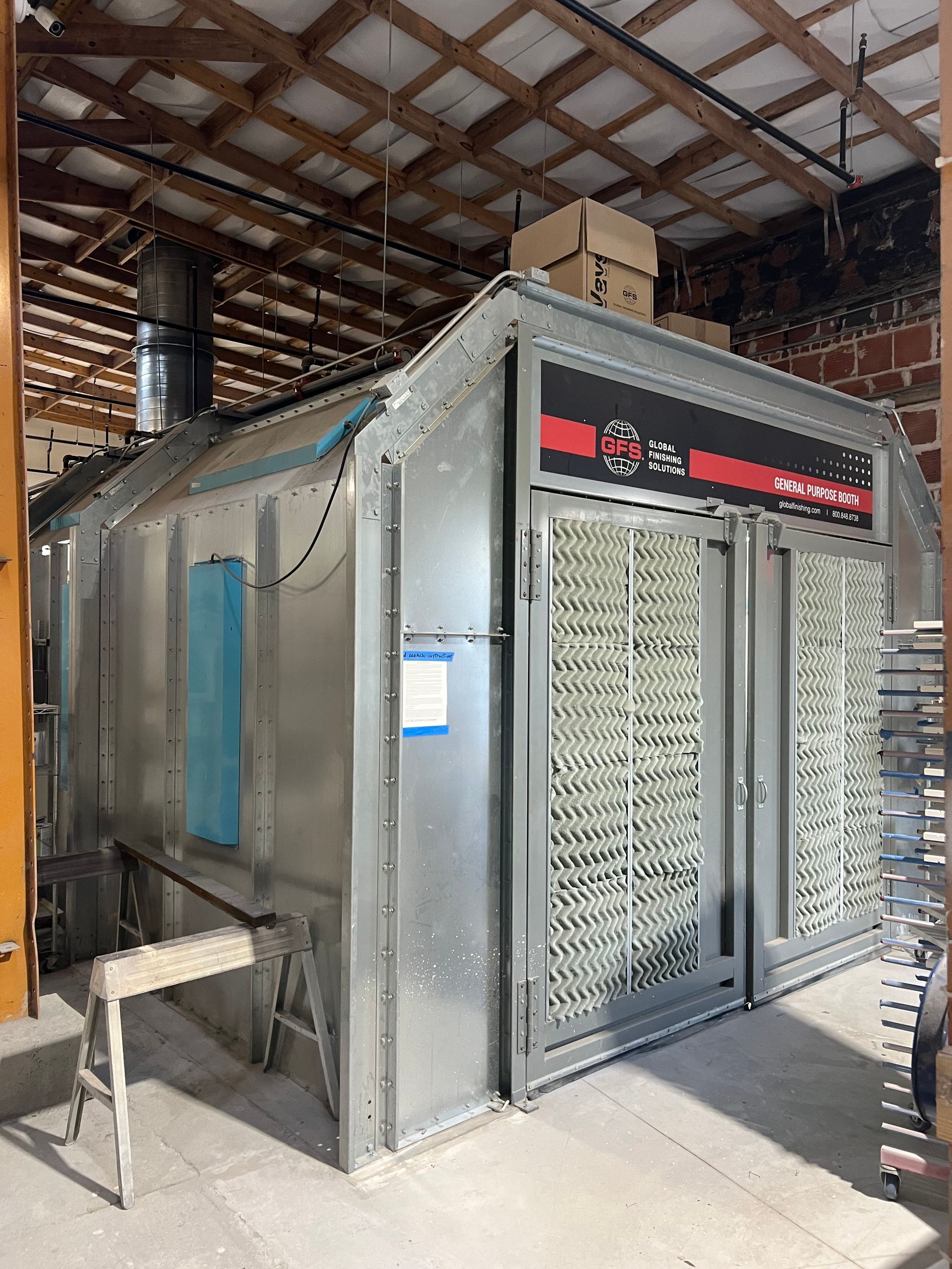 Metal paint booth in a workshop; it has a large front grate and a ventilation stack.