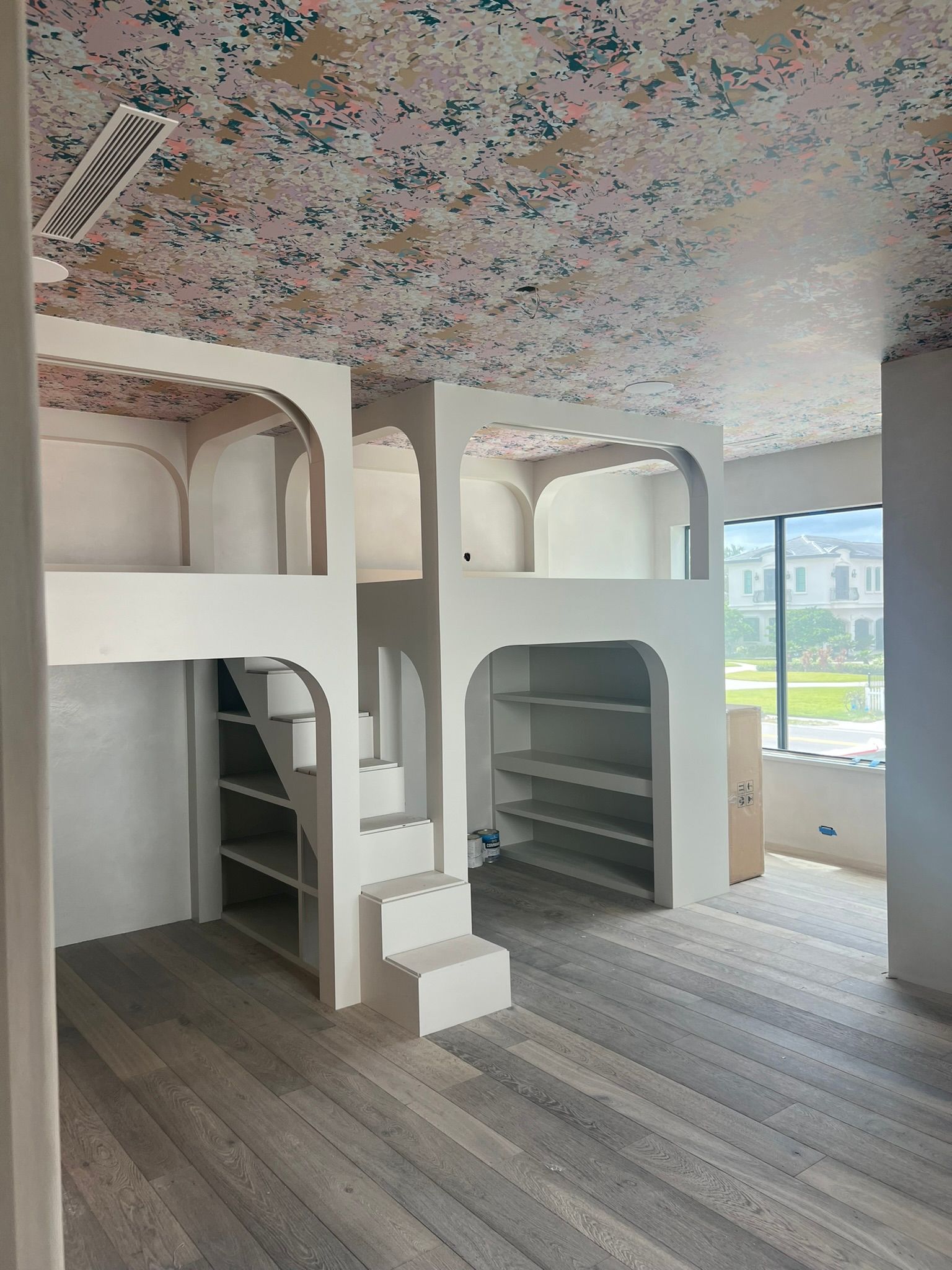 Room with built-in bunk beds and shelving. Floral wallpaper. Gray wood floor, white walls. Large window with a view.