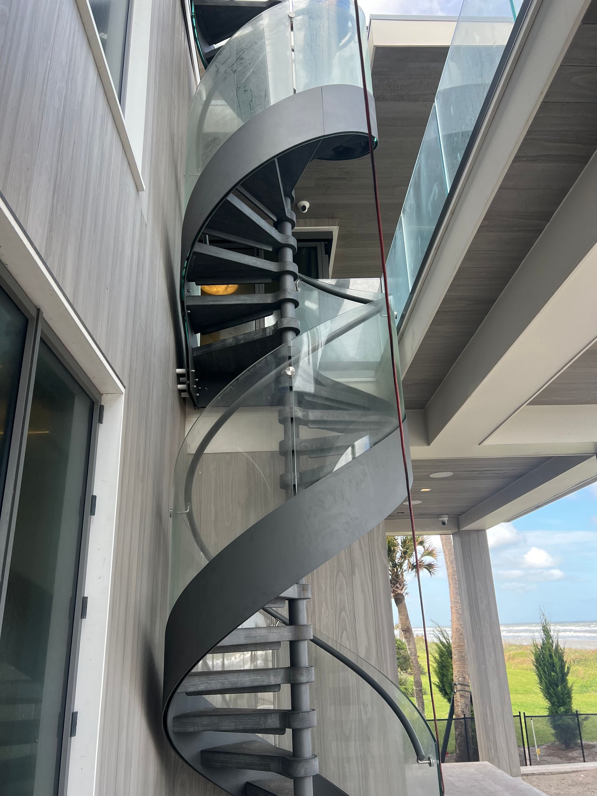Exterior gray spiral staircase with glass panels. Building exterior and beach visible.