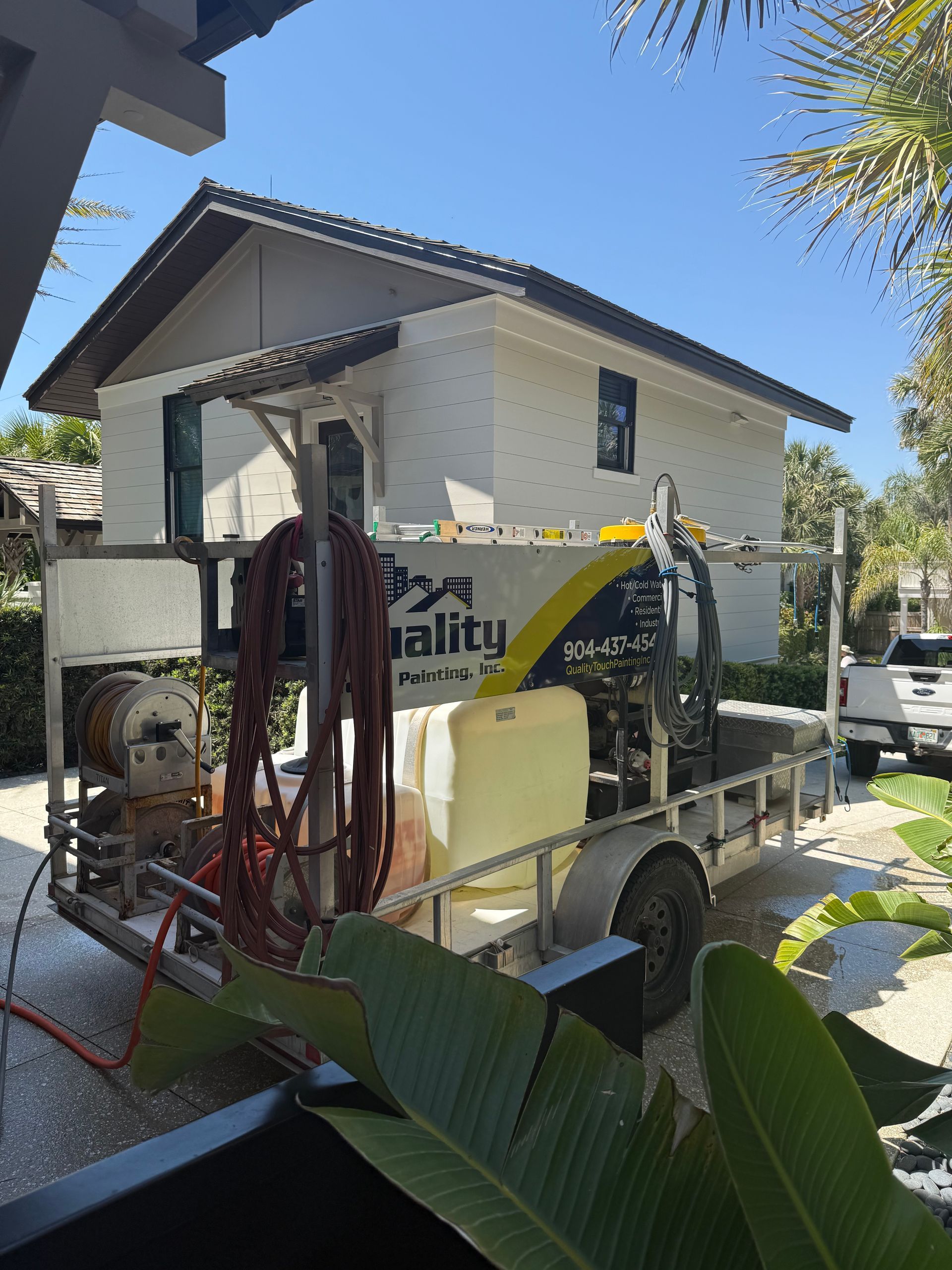 A trailer with pressure washing equipment parked near a white house with a brown roof.