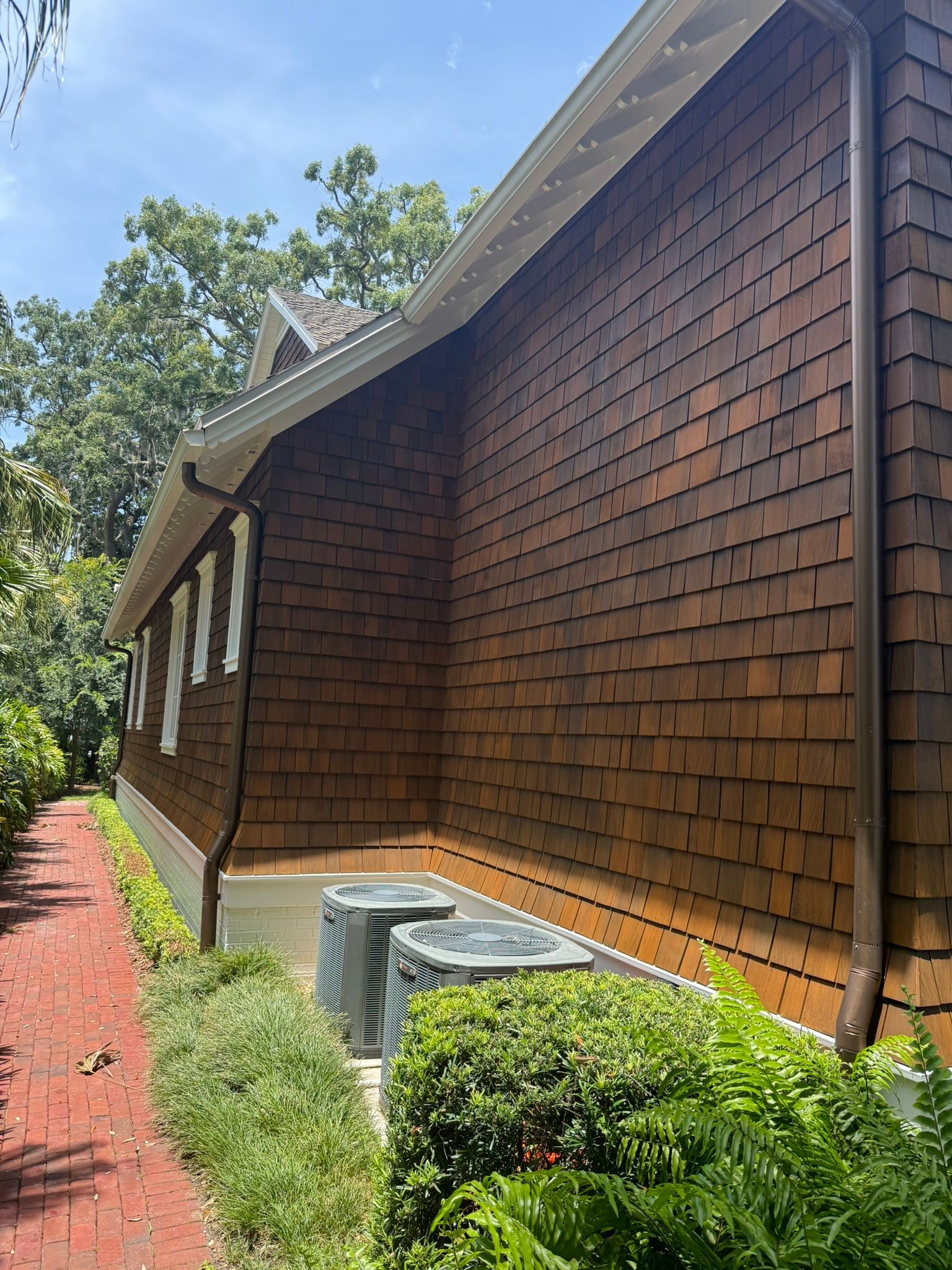 Brown shingled building with brown gutters, surrounded by greenery and a brick path.