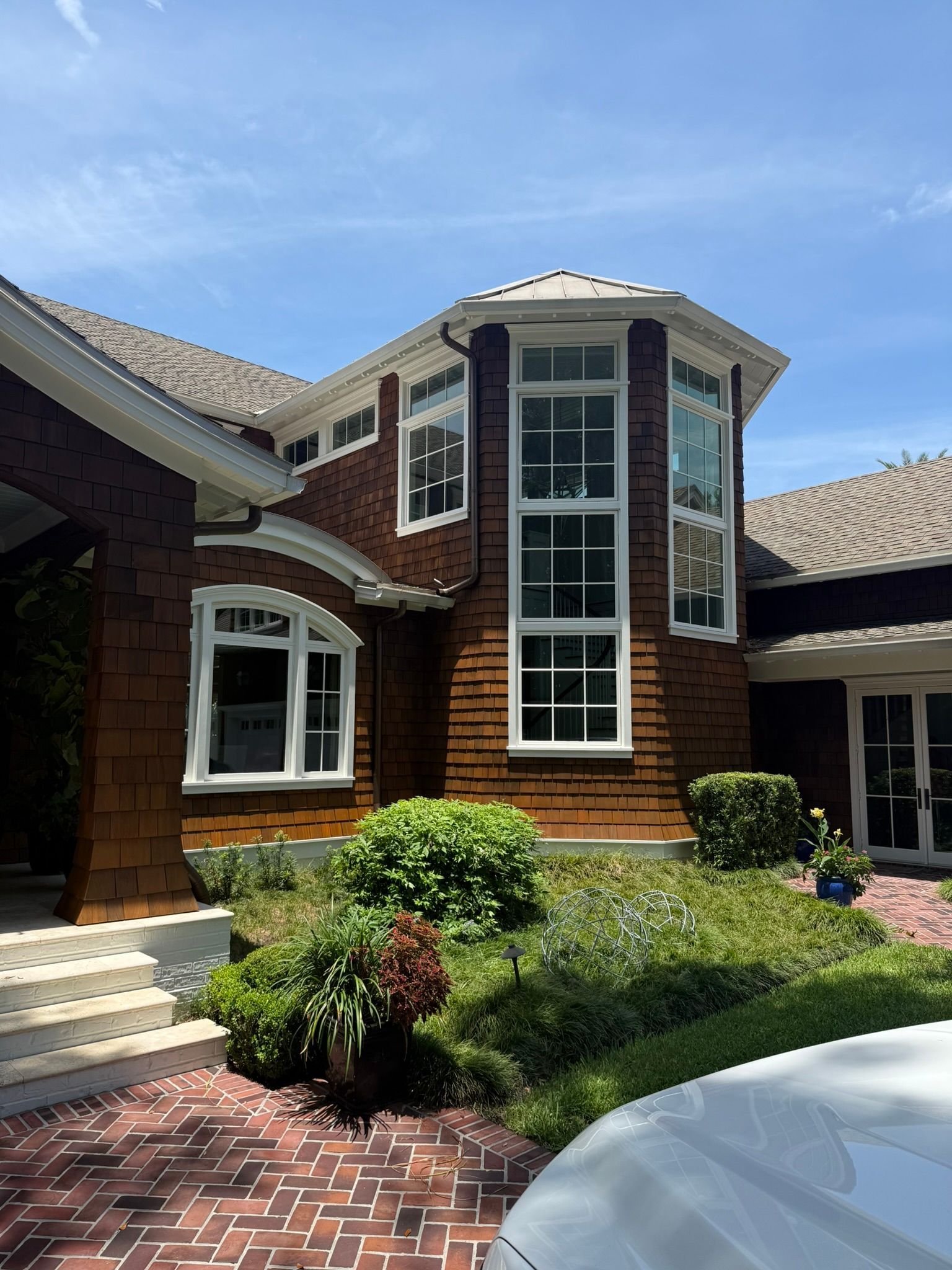 Brown shingled house with white-framed windows, brick driveway, and green landscaping under a blue sky.