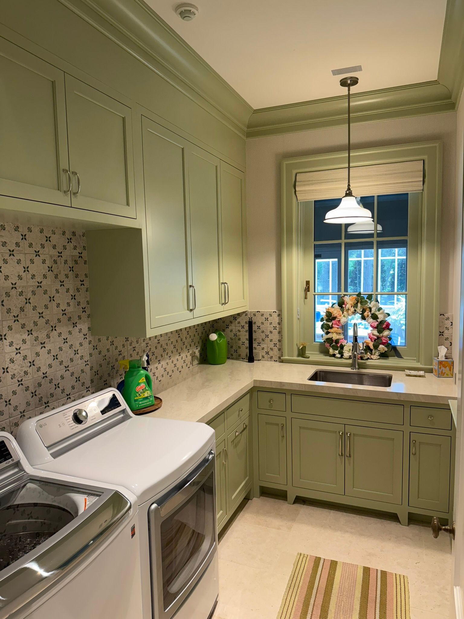 Laundry room with light green cabinets, white appliances, and window with wreath.