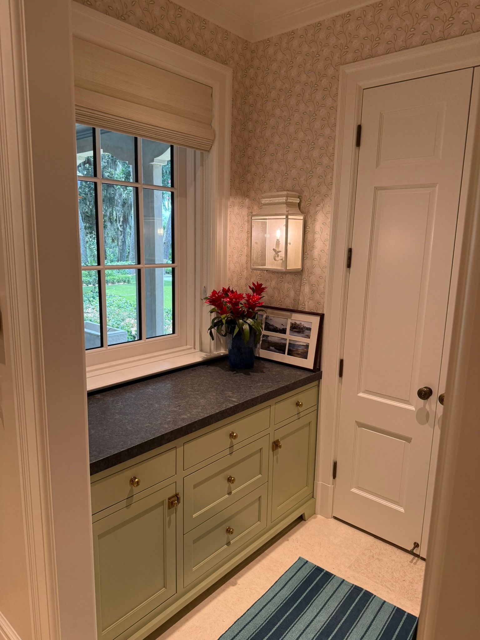 Green cabinets with black countertop under a window, red flowers, a framed picture, a door, and a lantern.