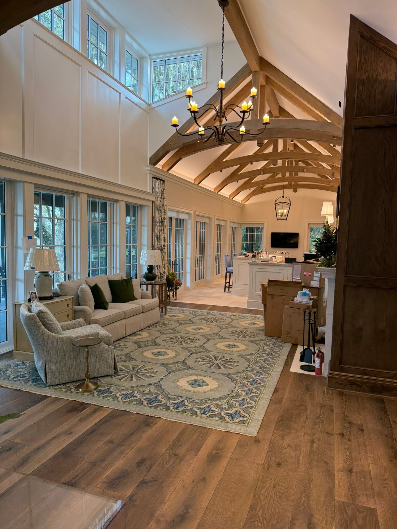 Living room with high ceilings, wood beams, and large windows.  Sofa, rug, and chandelier present.