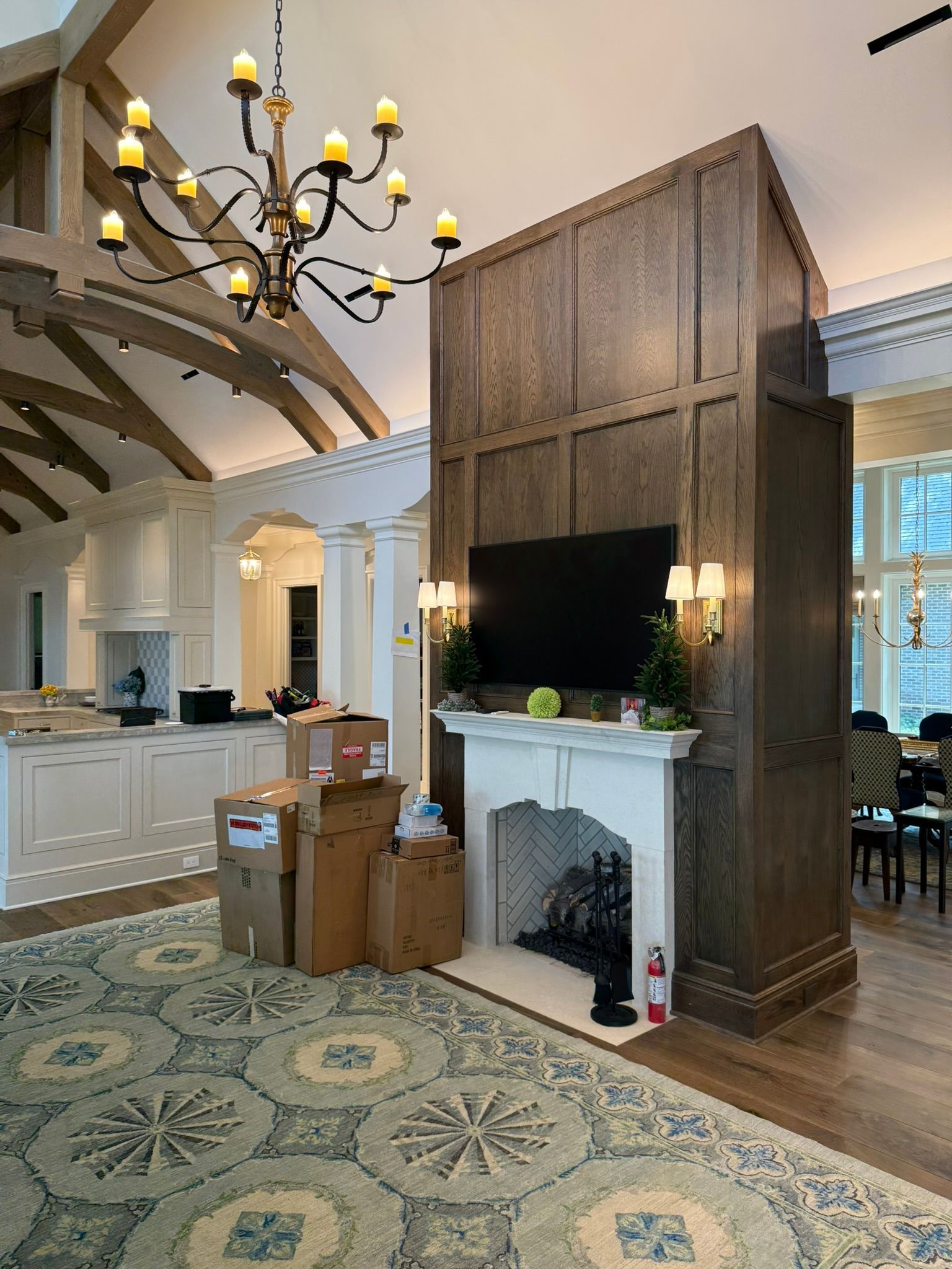 Living room with fireplace, TV, and boxes.  A chandelier hangs overhead. Light wood and patterned rug.