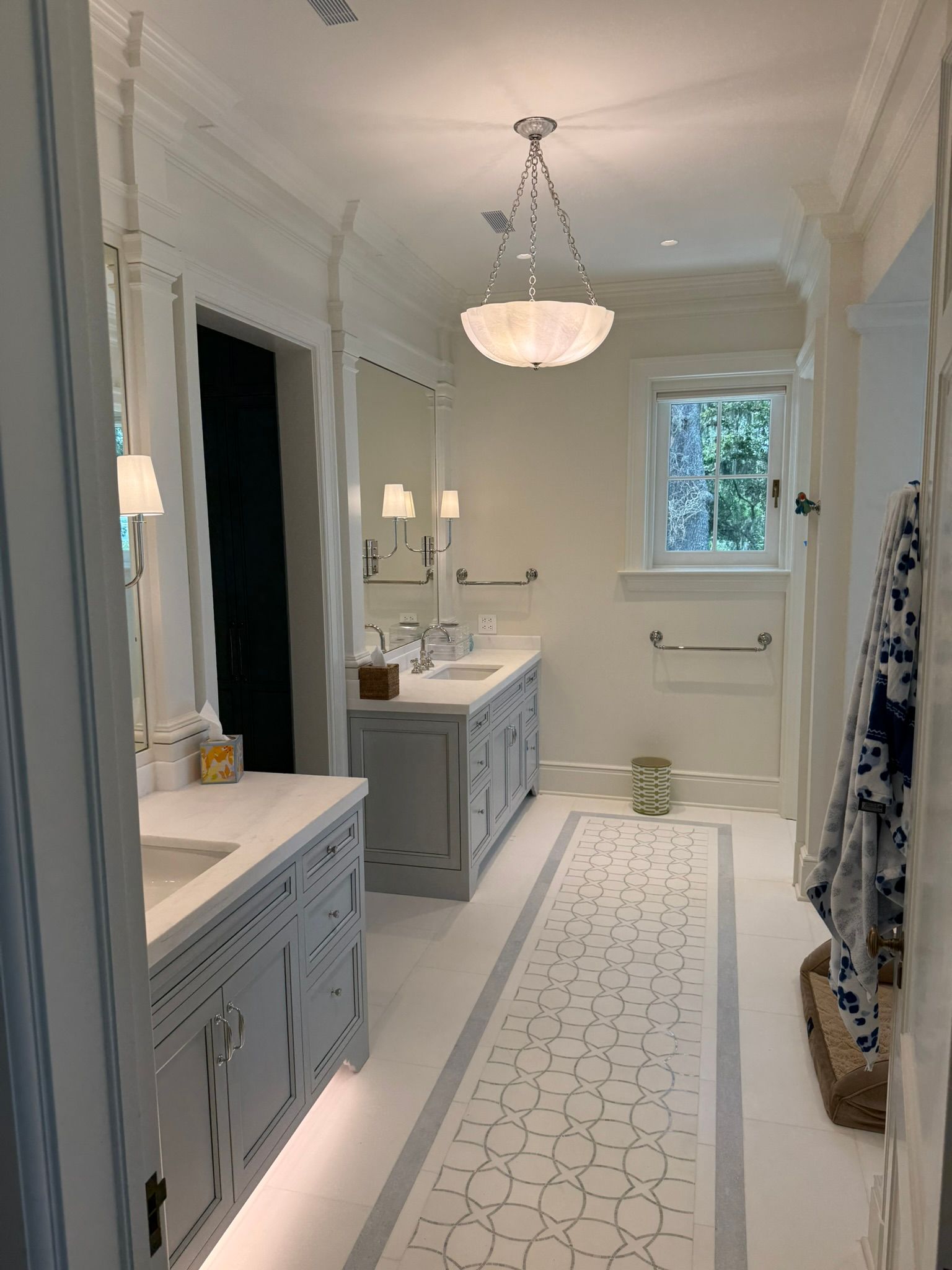 Elegant bathroom with gray vanities, white tile floor with mosaic runner, and chandelier.