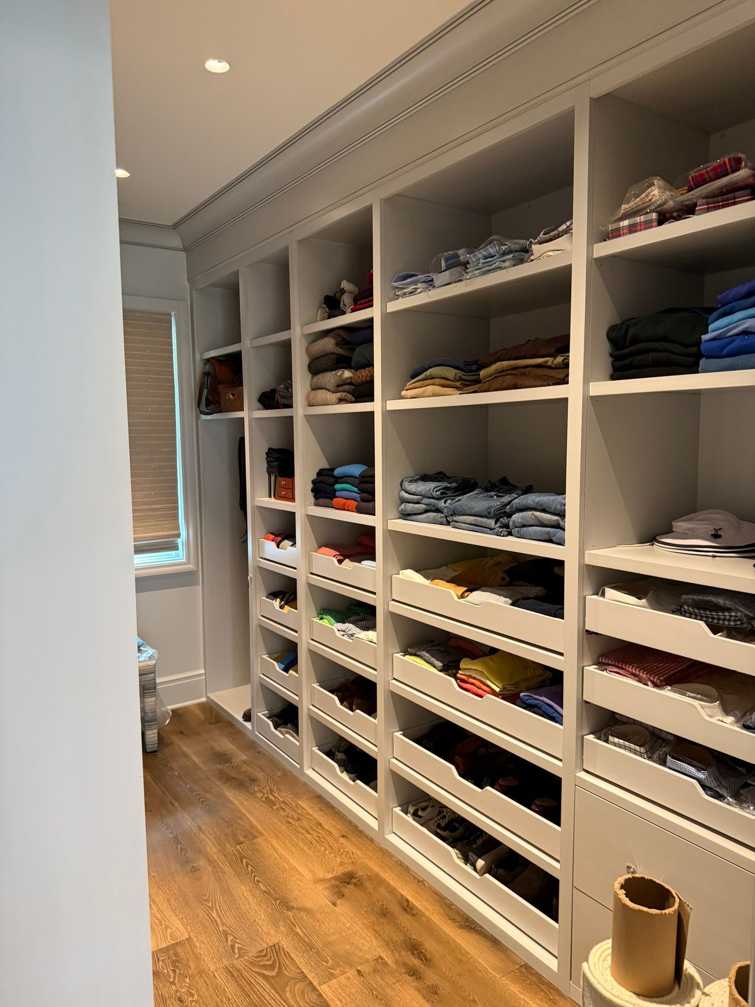 Built-in, white closet with shelves holding folded clothes. Wood floor, window with blinds on left.