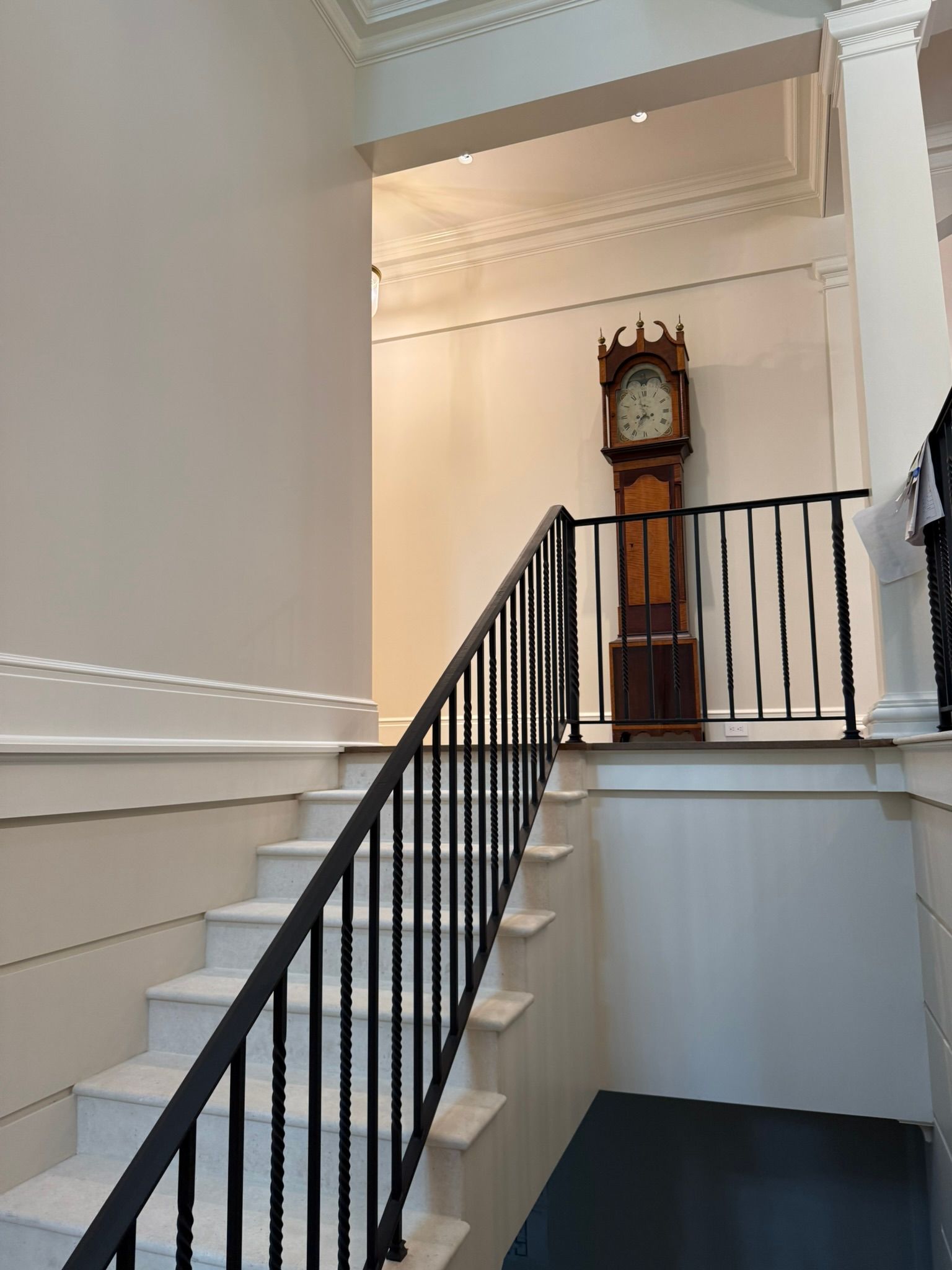 Staircase with black railing leading up to an antique grandfather clock. White walls, cream trim.
