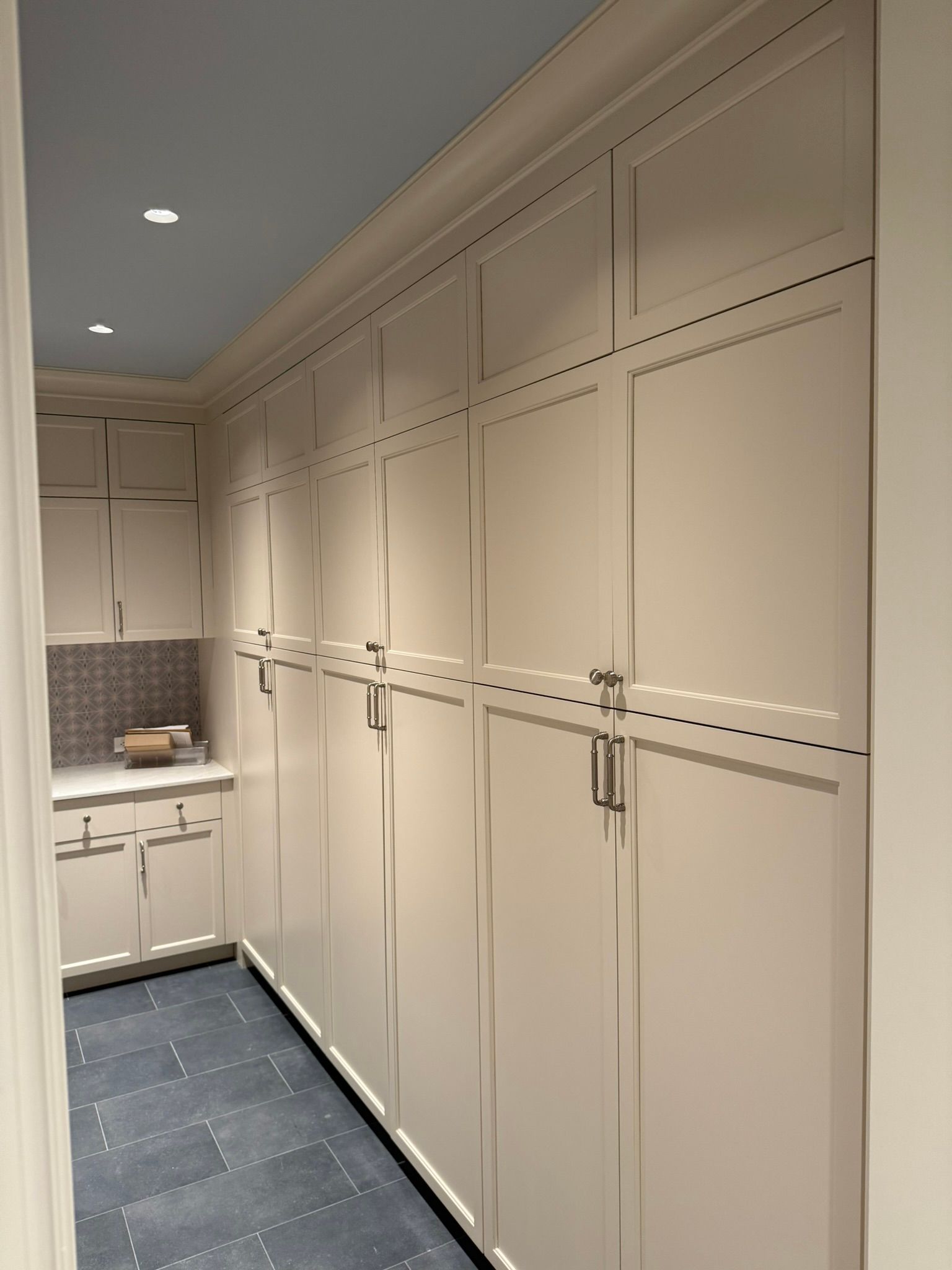 Long, white, built-in storage cabinets line a hallway, with a small counter area and blue floor tiles visible.