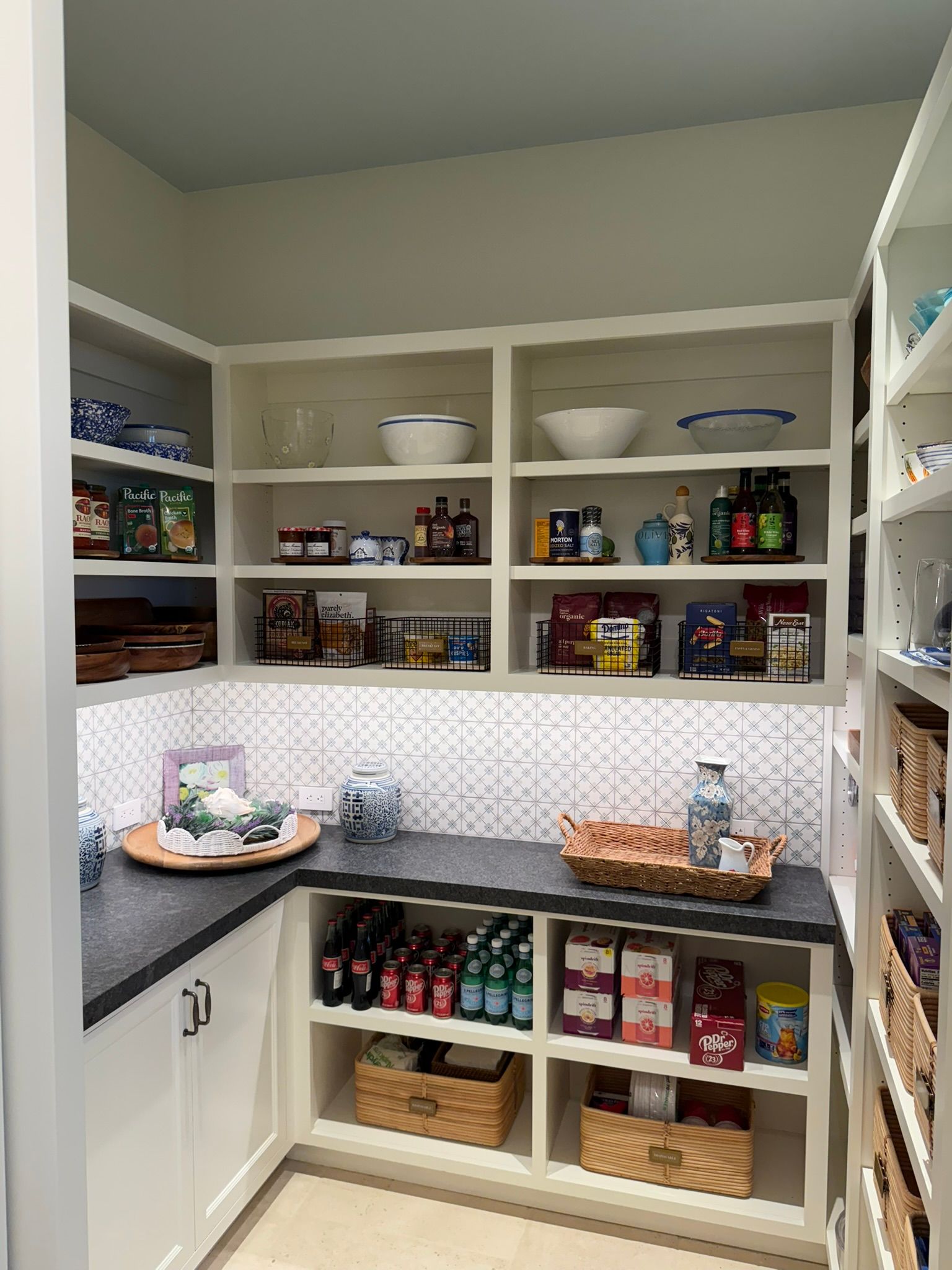A well-organized pantry with white shelves, black countertops, and various food items and baskets.