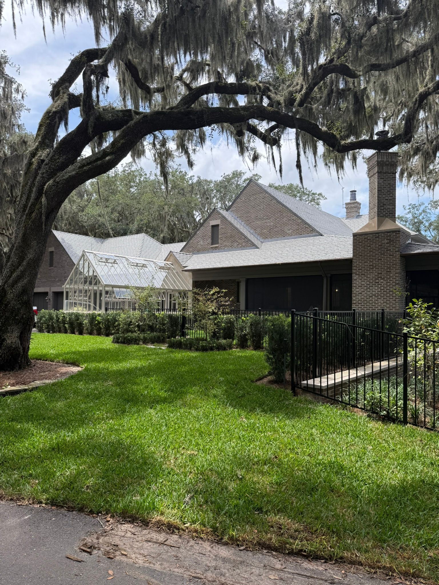 Lush green lawn with house under large tree. Light-colored metal roof, stone chimney, and black fence.