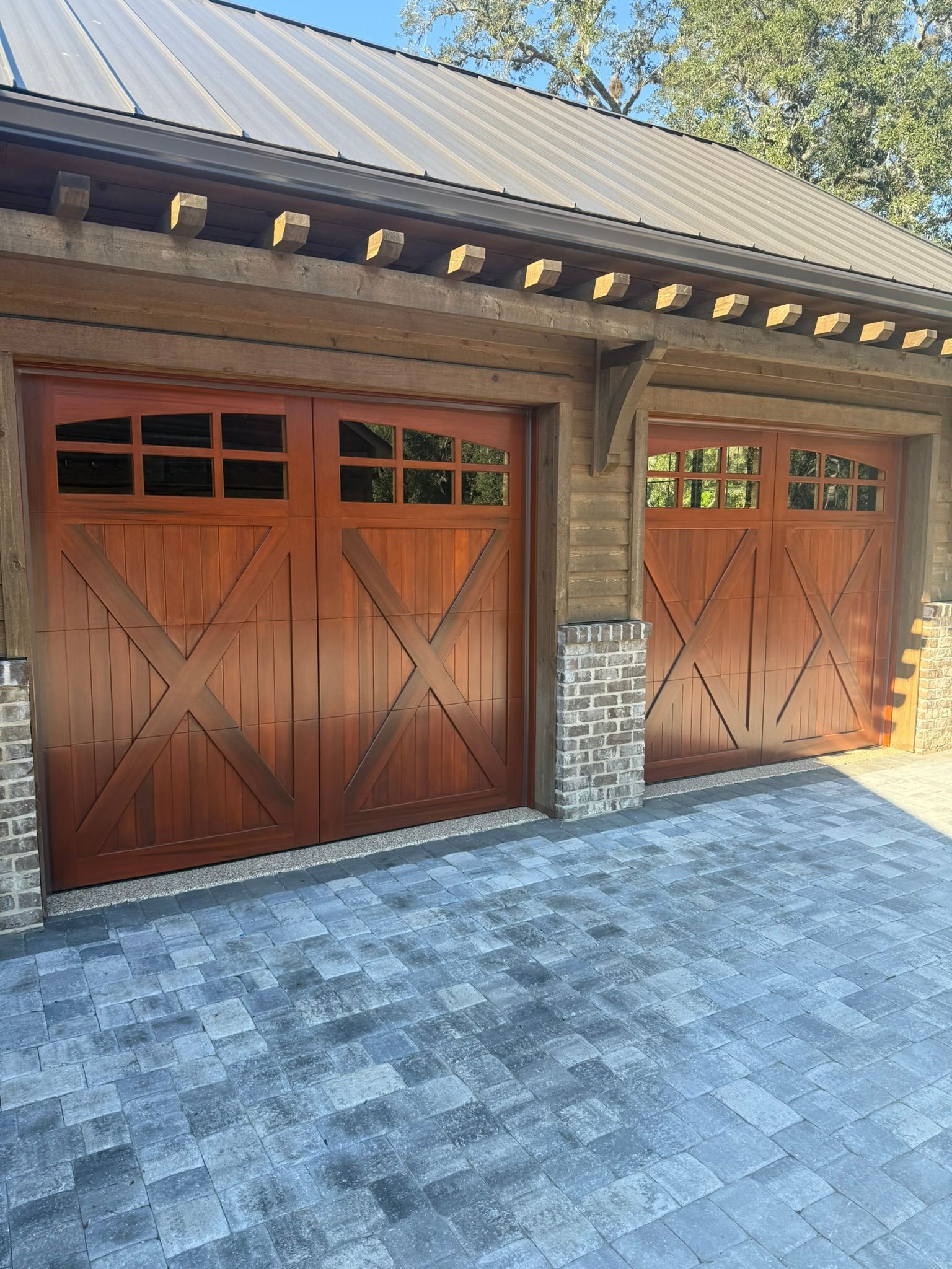 Two brown wooden garage doors with cross design, brick accents, and a paved driveway.