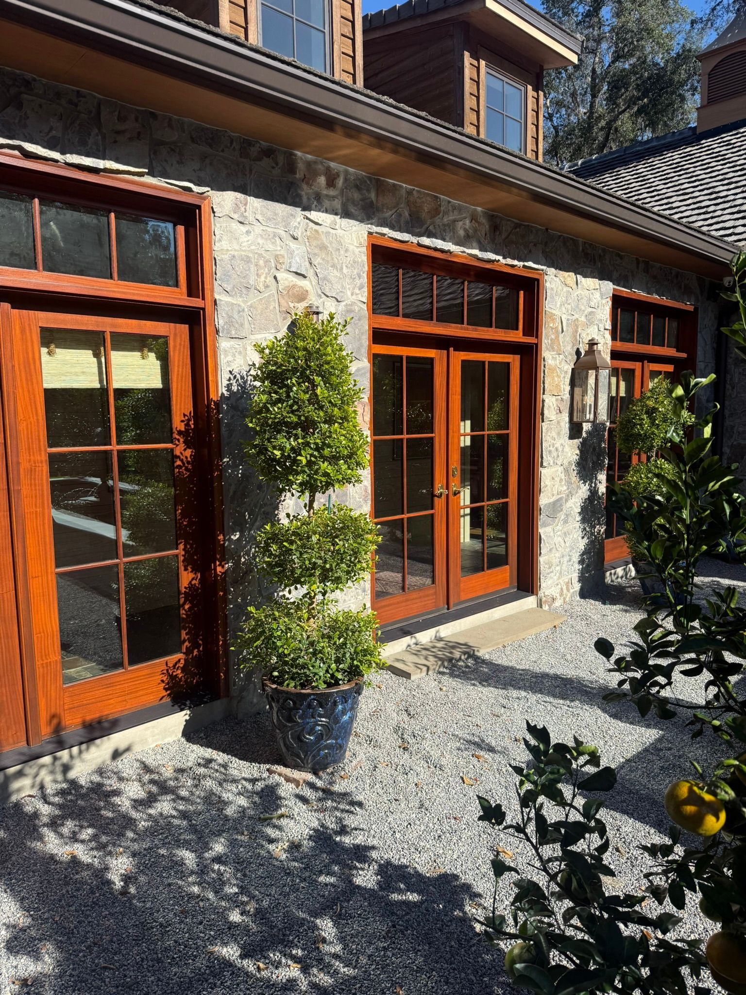 Stone building with brown wood doors and windows, gravel patio, and potted trees.