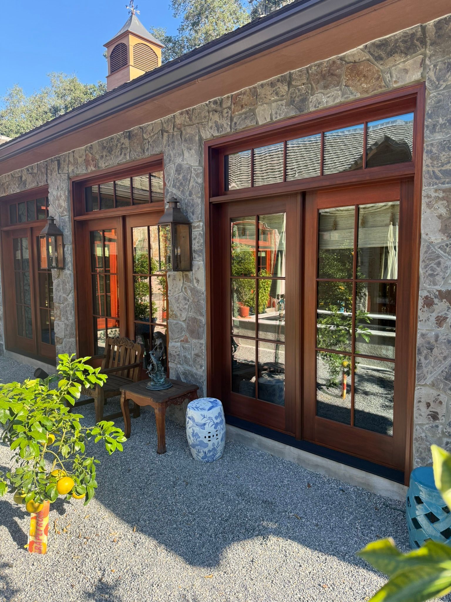 Exterior view of a stone building with wooden-framed doors reflecting a garden, sitting area, and blue and white ceramic stool.
