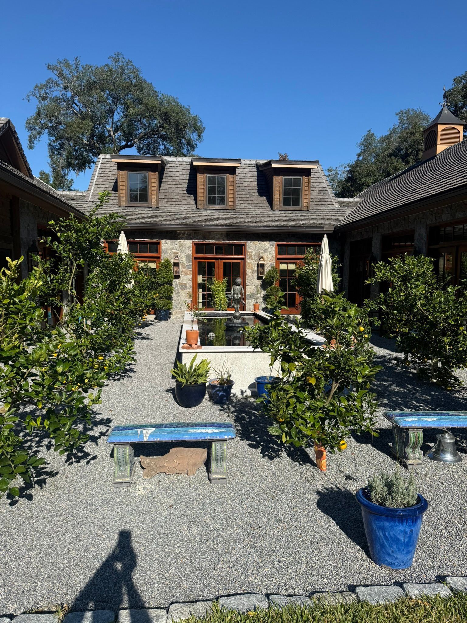 Courtyard with trees, blue benches, and a stone building with a slate roof under a bright blue sky.