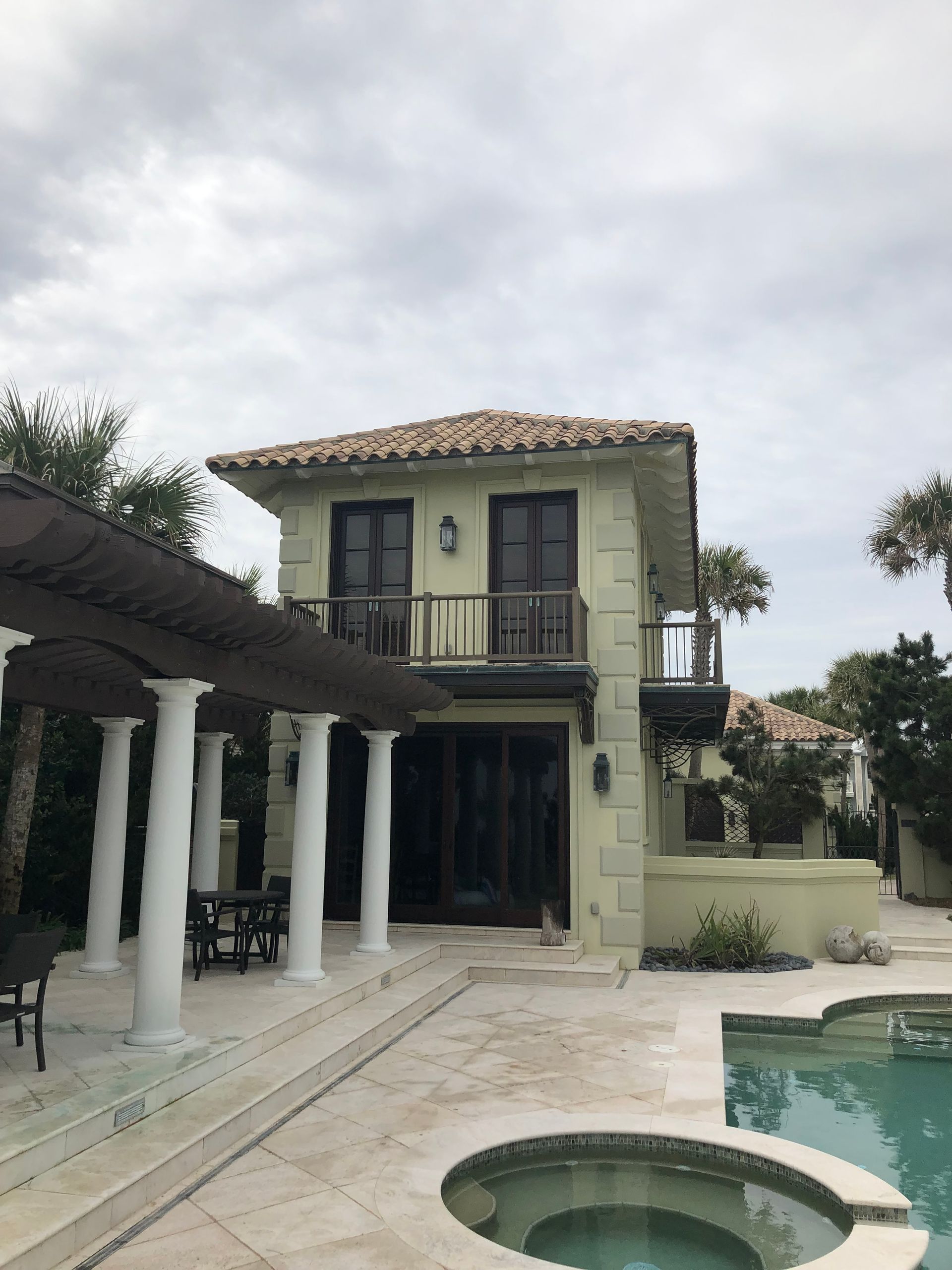 Two-story Mediterranean-style house with pool and pergola; beige exterior, tile roof, and cloudy sky.
