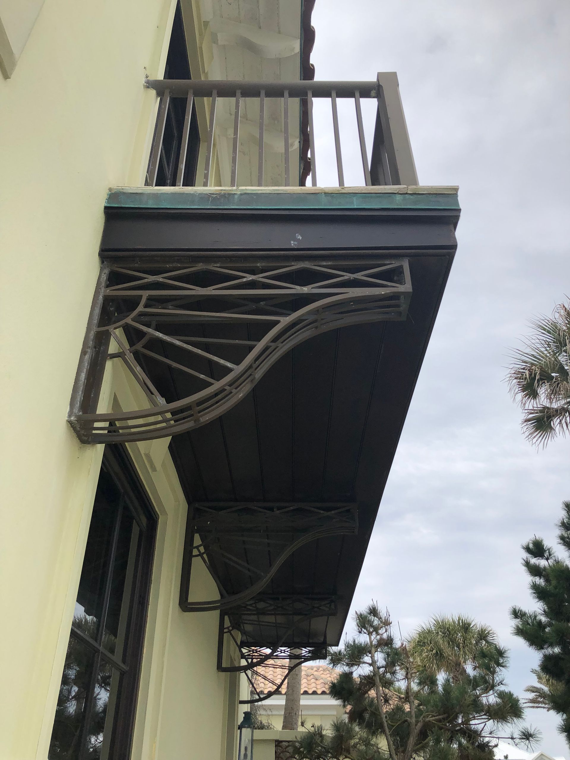 Balcony with ornate wrought iron supports, brown railing, and dark underside, attached to a cream-colored building.