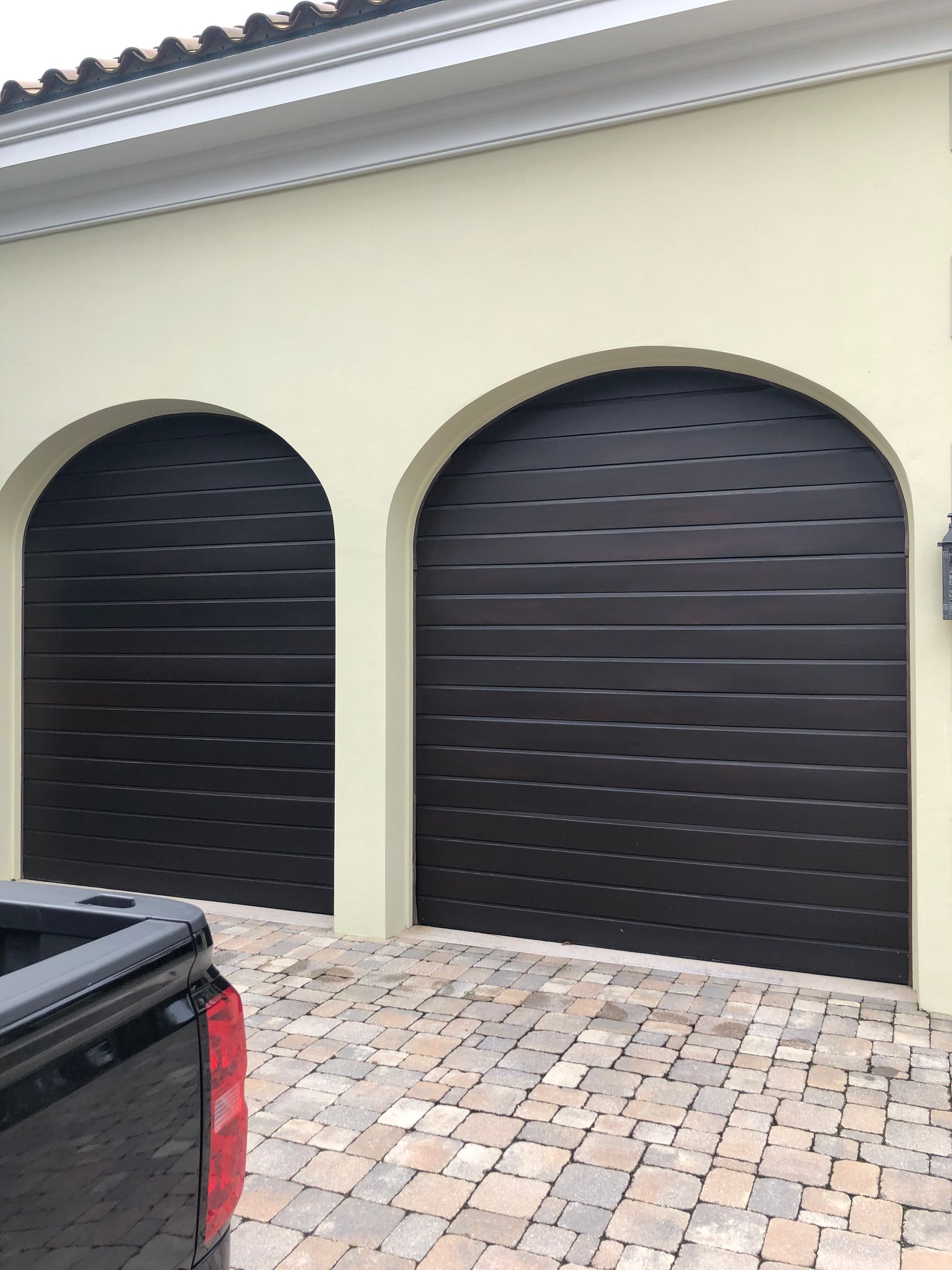 Two arched black garage doors on a light yellow building, cobblestone driveway, partial view of a black truck.