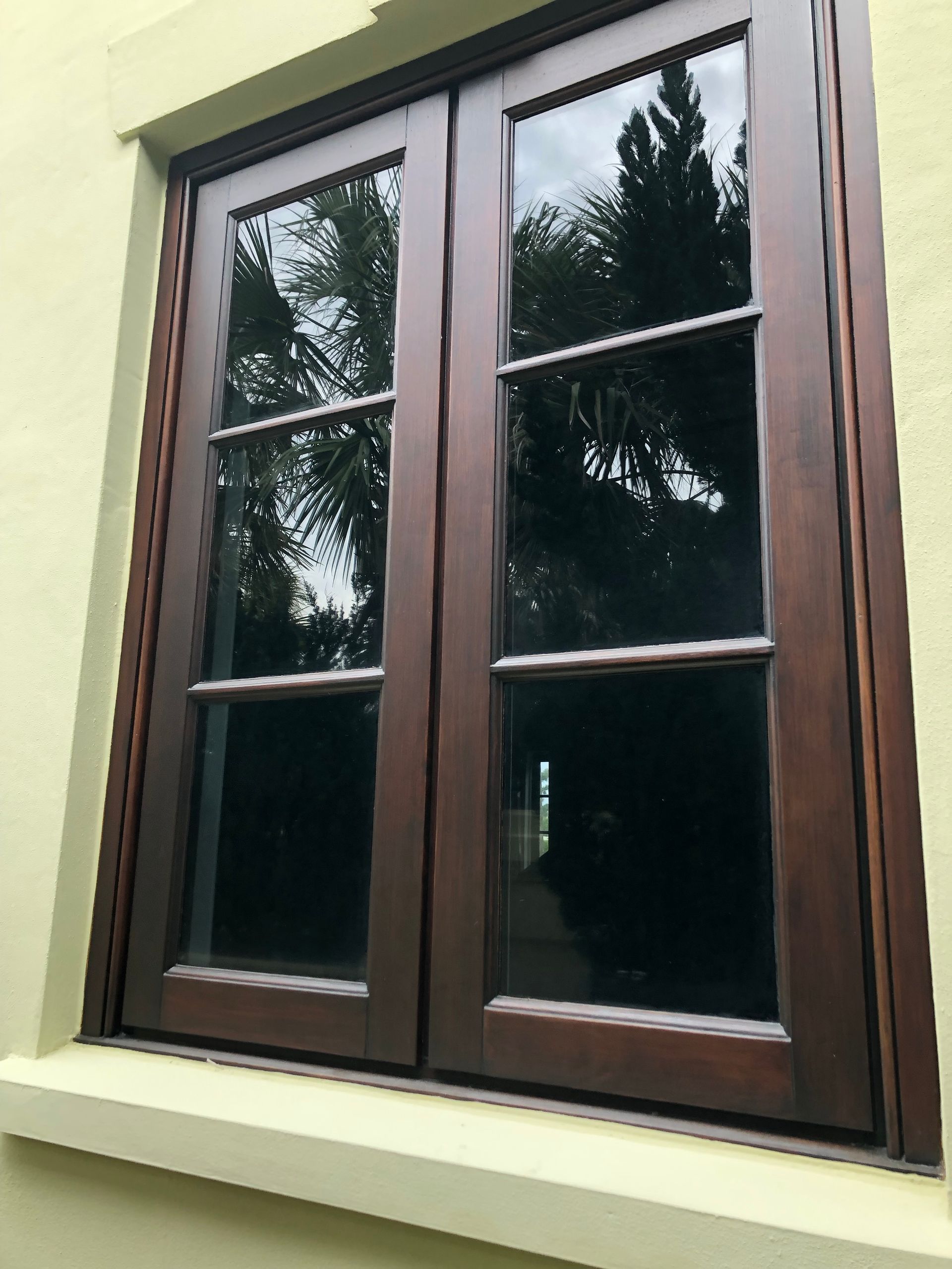 Dark wooden double window with clear glass panes reflecting outdoor trees, set in a light yellow wall.
