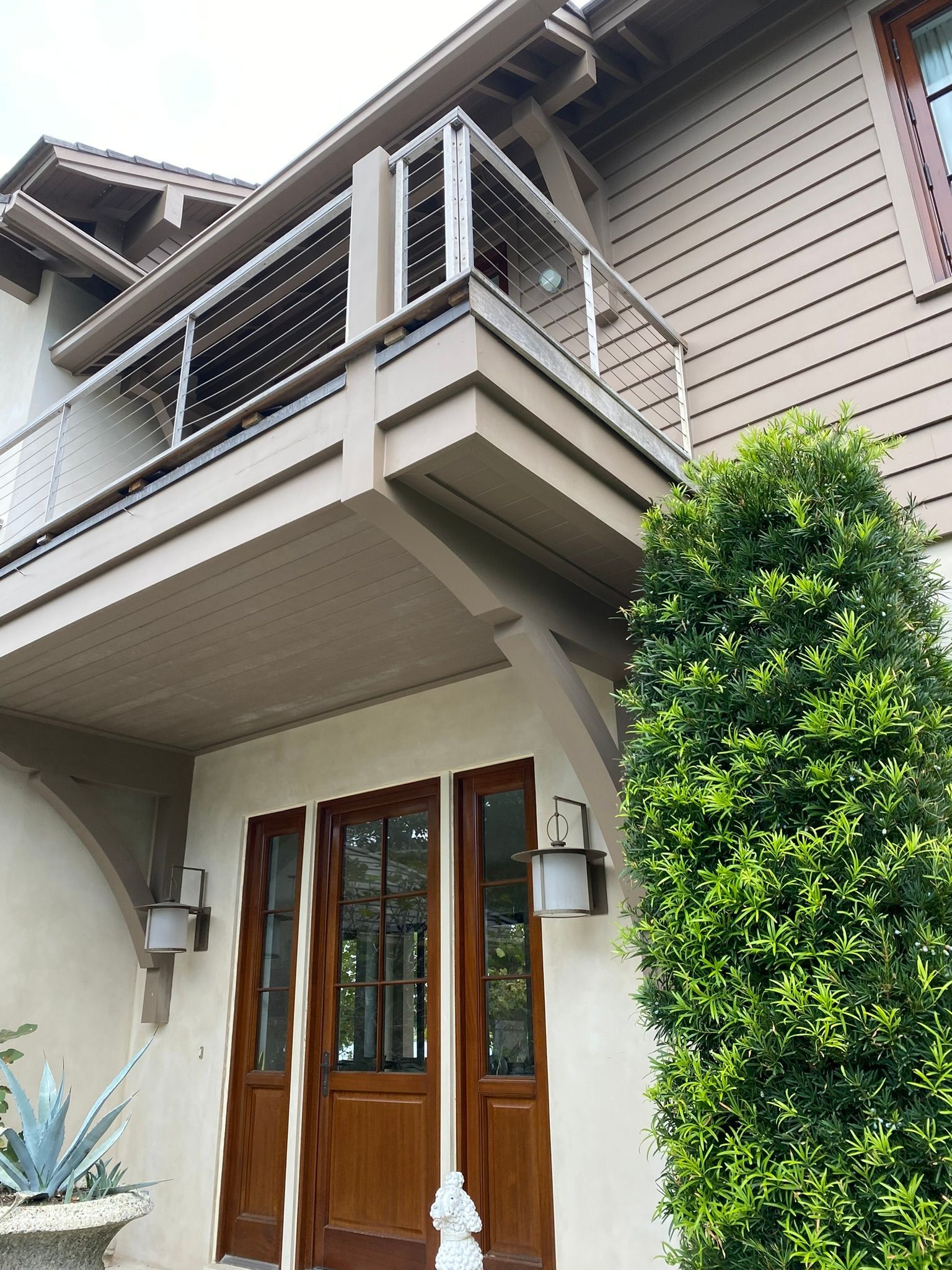 Beige house with a balcony and dark wooden doors. A green bush is to the right.