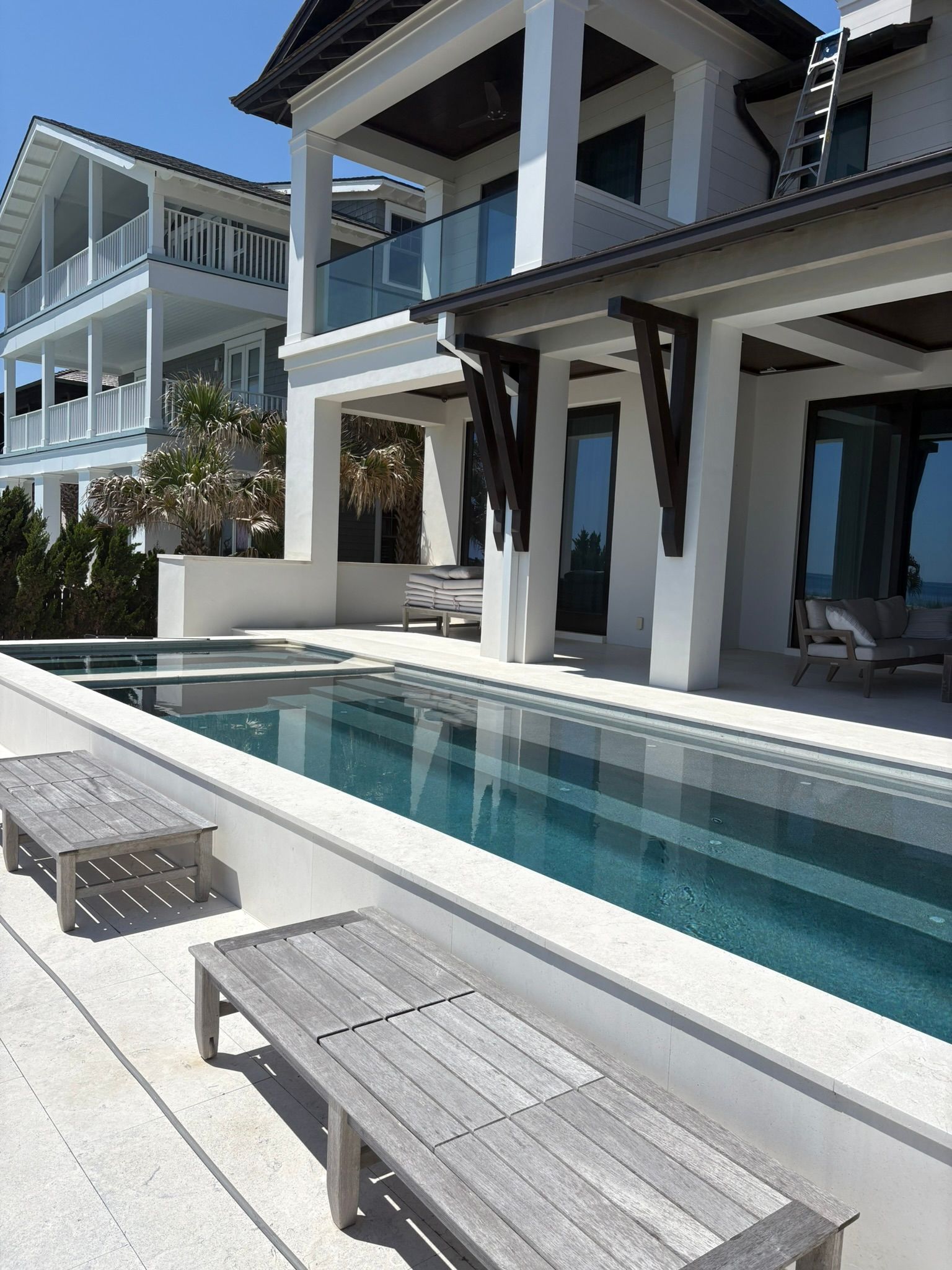 Luxurious white house with a long pool and wooden benches, sunny outdoor setting.
