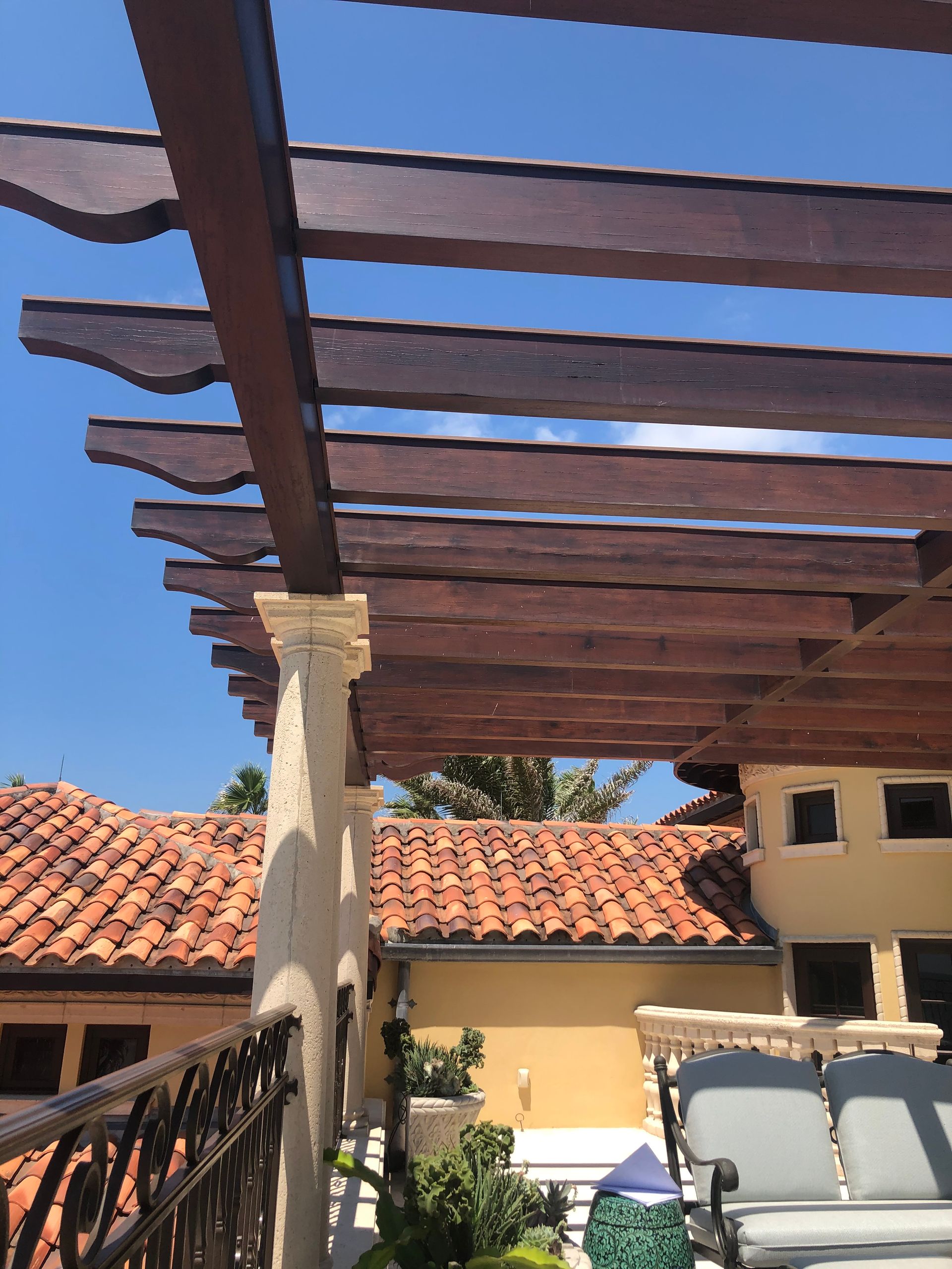 Pergola with dark wood beams over a terracotta roof, supported by a beige pillar; blue sky background.