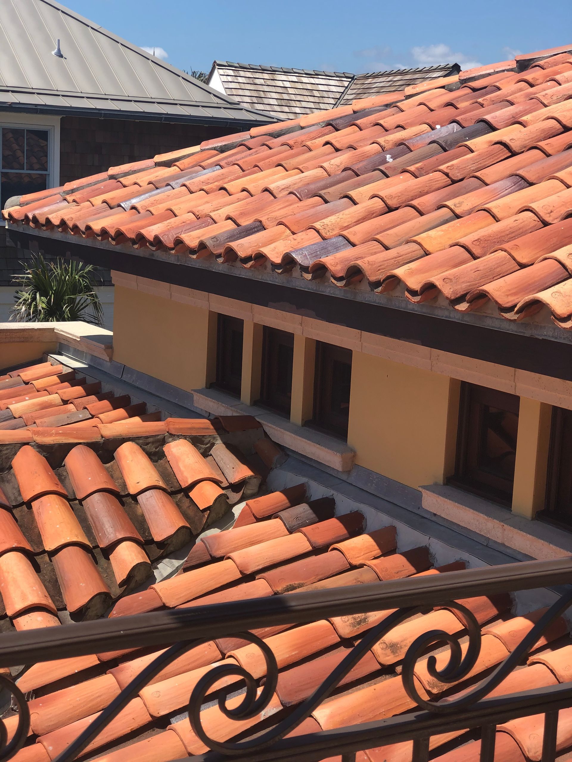 Terracotta tile roofs on buildings, with a wrought-iron balcony railing in view. Blue sky.