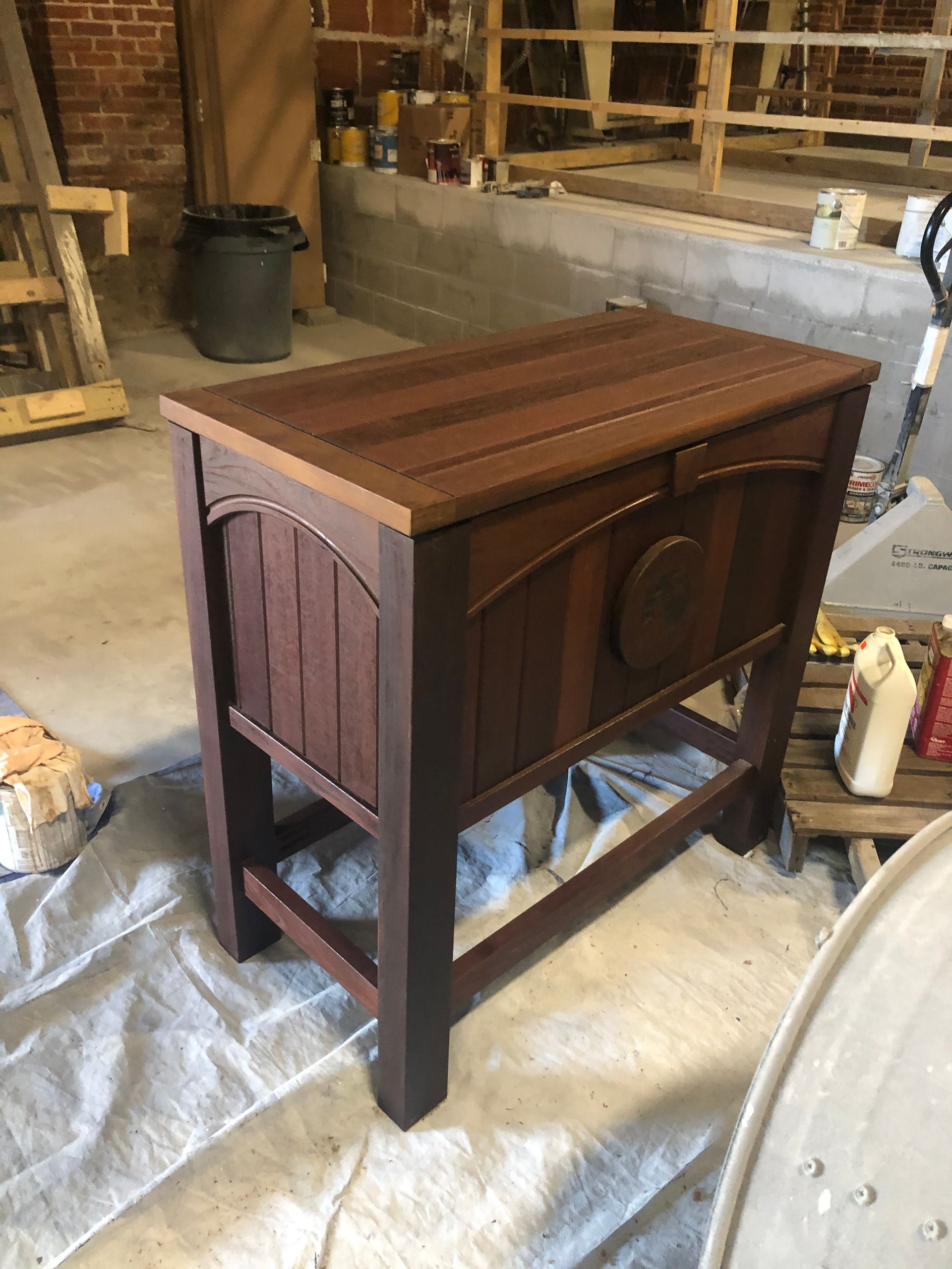 Wooden cooler chest with arched paneling and a dark stain, on a stand in a workshop.