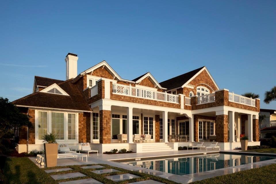 Large house with brown shingle siding, white columns, balcony, and a swimming pool reflecting the blue sky.