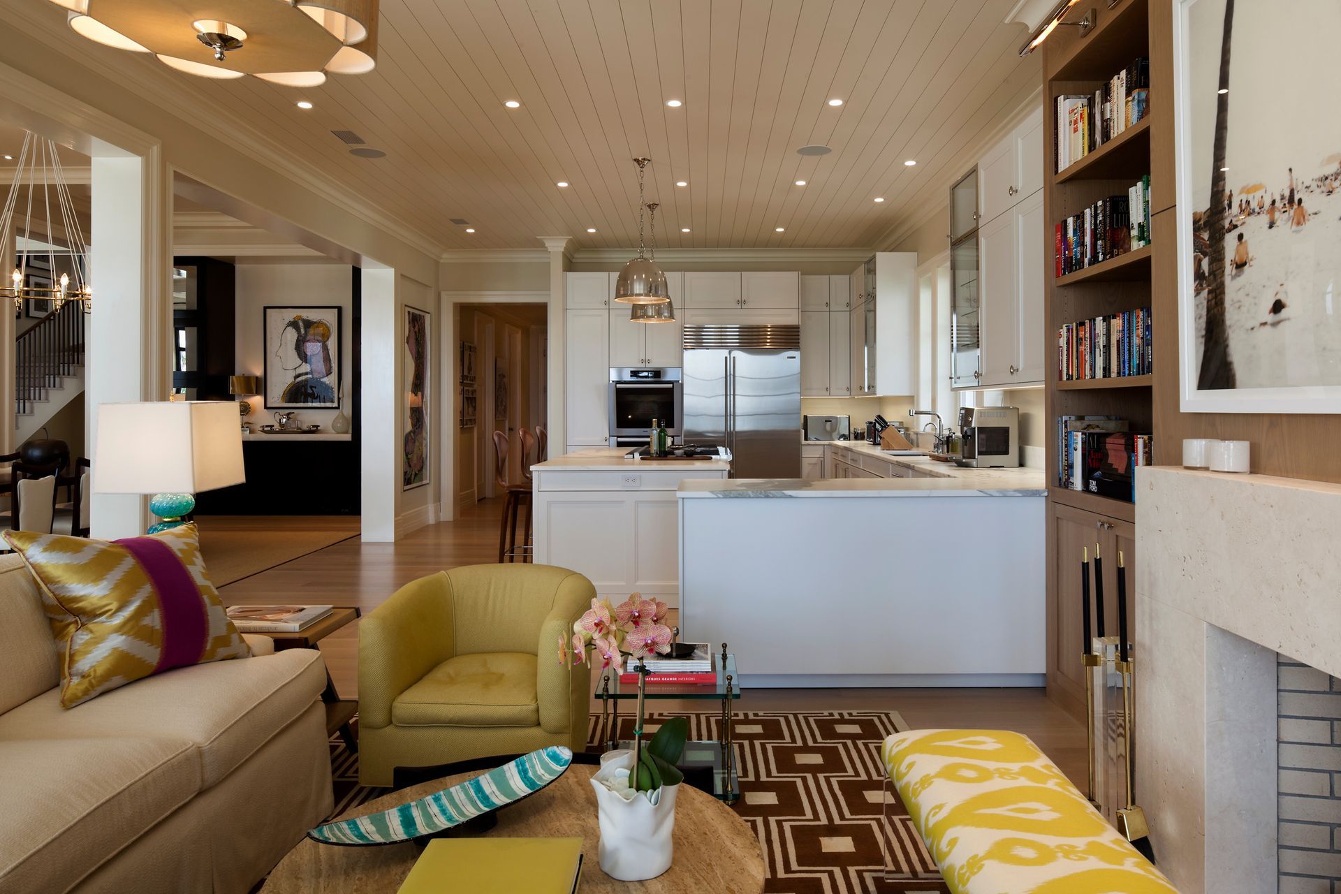 Living room with kitchen in the background; cream and yellow furniture, white walls, wood floors, and a patterned rug.