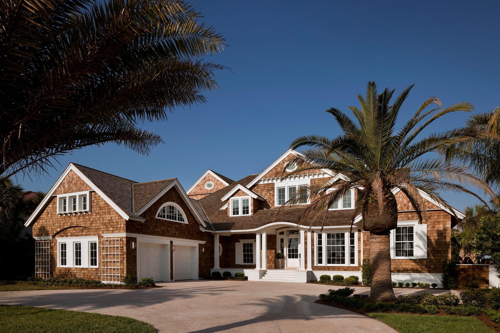 Beachfront house with wood siding, brown roof, and palm trees. Driveway leads to the garage.