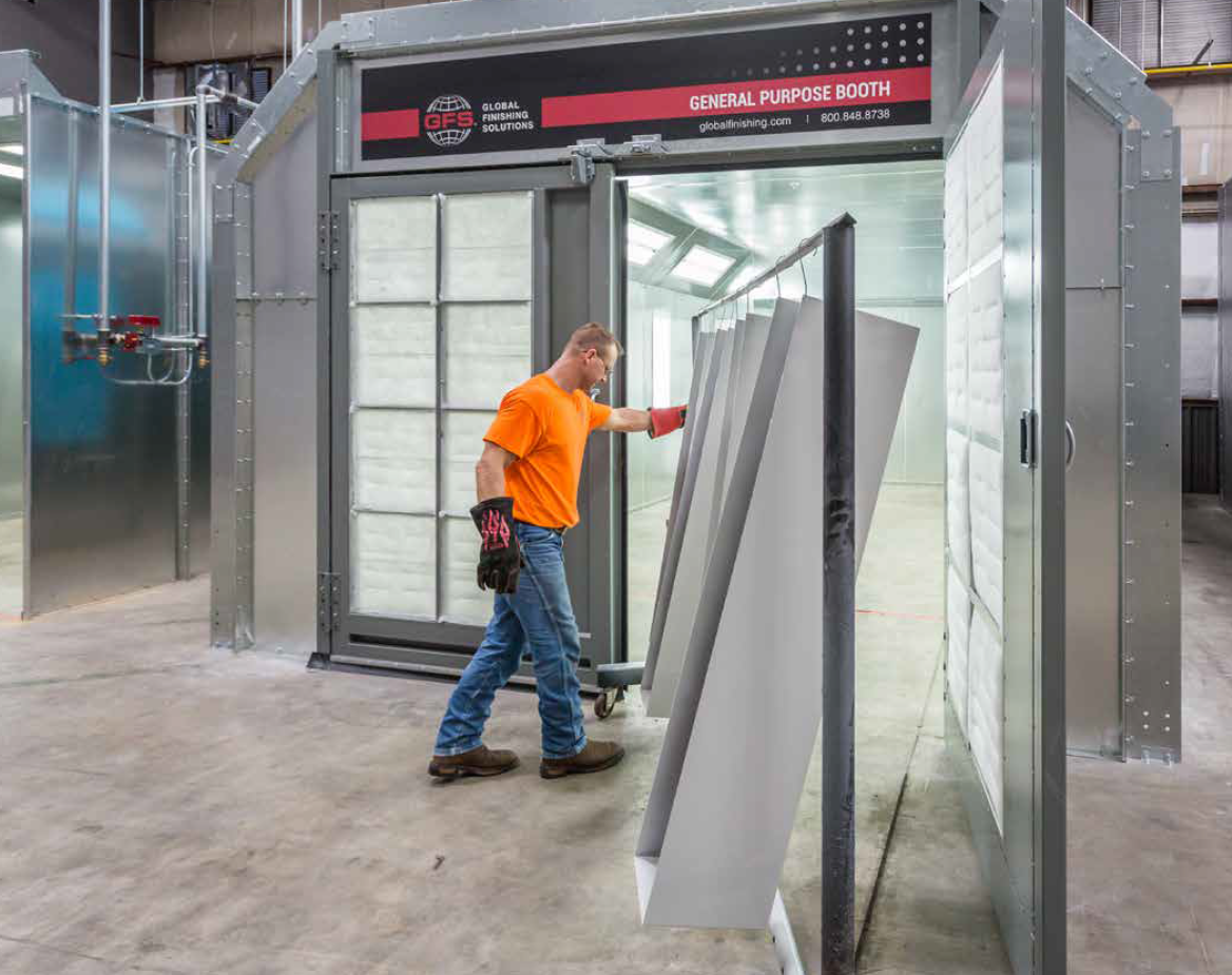 Man in orange shirt placing metal panels in a paint booth, in an industrial setting.