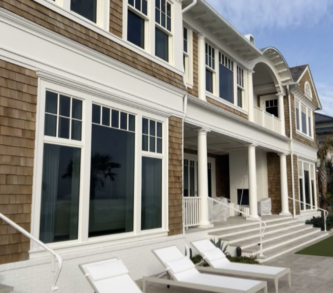 Large, light-colored house with tan shingles, white trim, and a porch. Several lounge chairs sit outside on a sunny day.