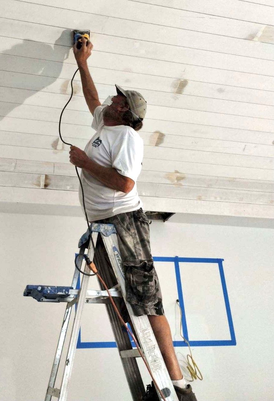 Man on a ladder sanding a white plank ceiling. He's wearing a hat and shorts; blue tape outlines a window.