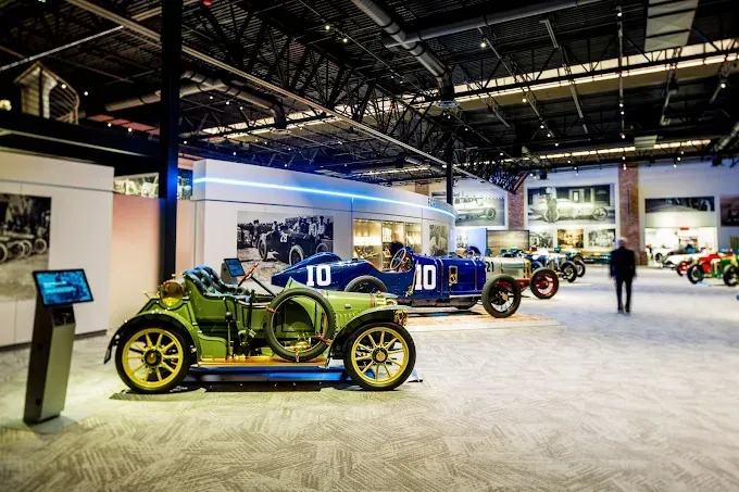 Classic cars on display in a museum. A person walks between the vehicles, with information screens visible.