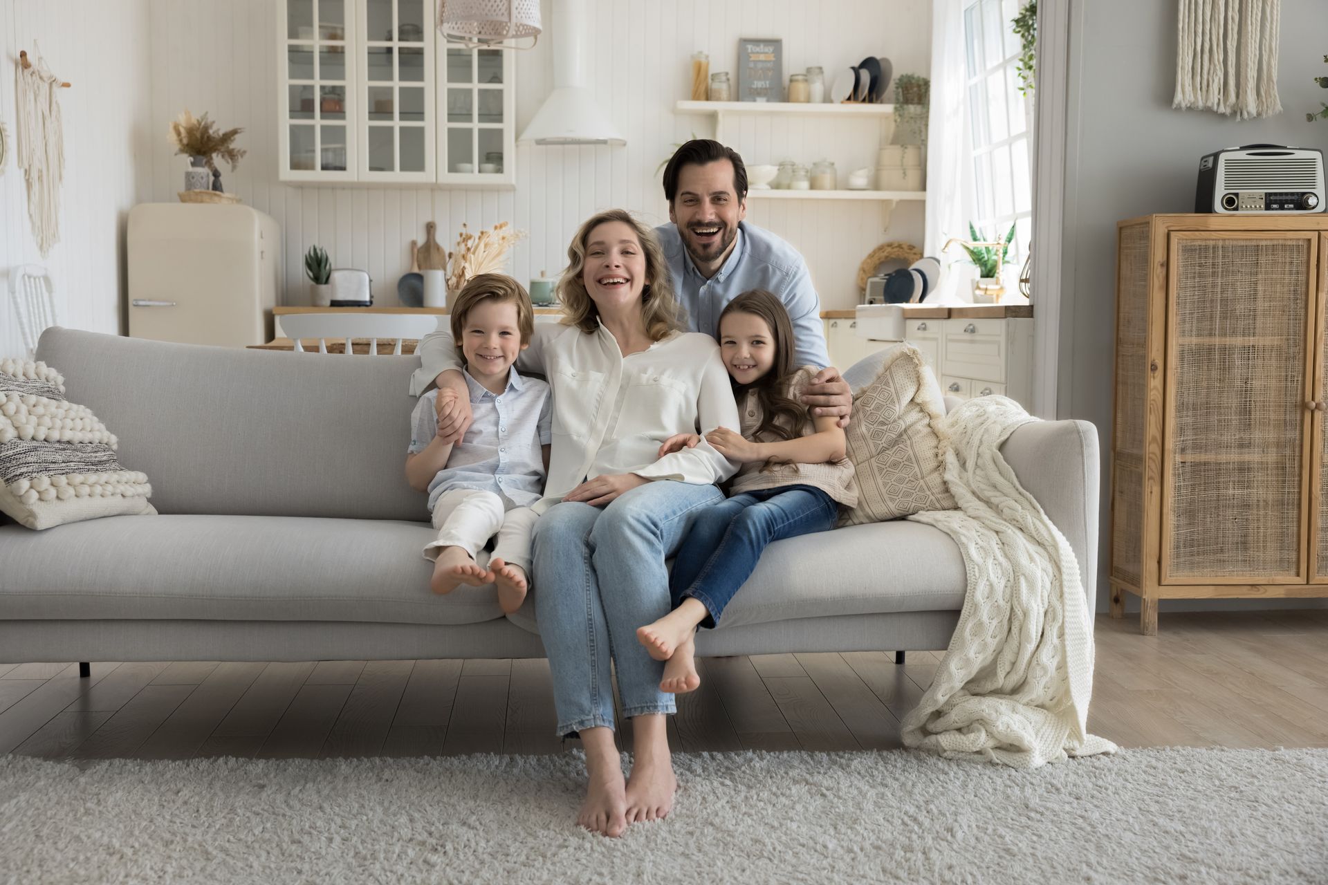 Family of four smiling, sitting on a light grey sofa in a bright living room, with a kitchen visible in the background.