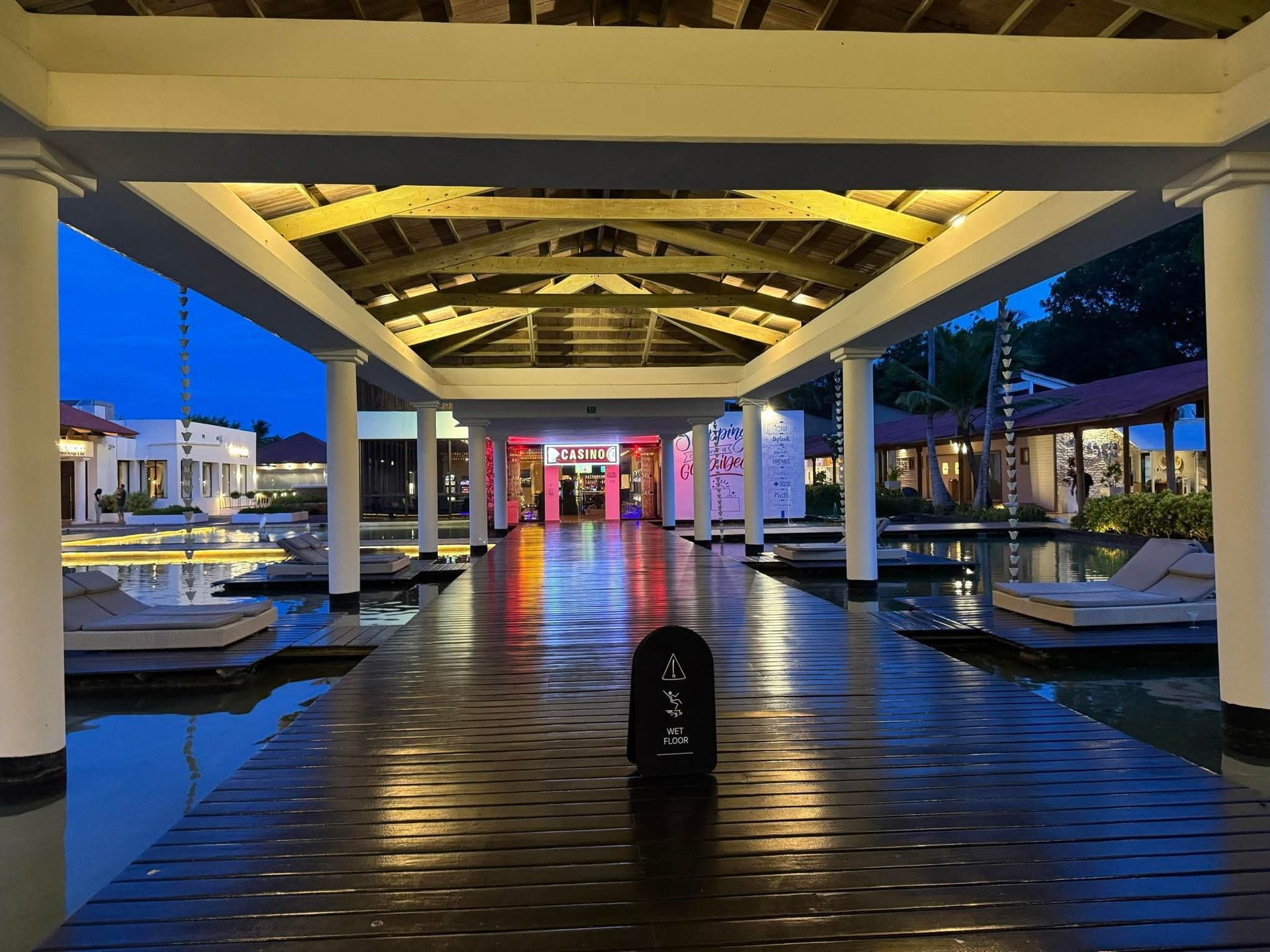 Long wooden walkway over water, flanked by lounge chairs. Buildings and colorful lighting in the distance.