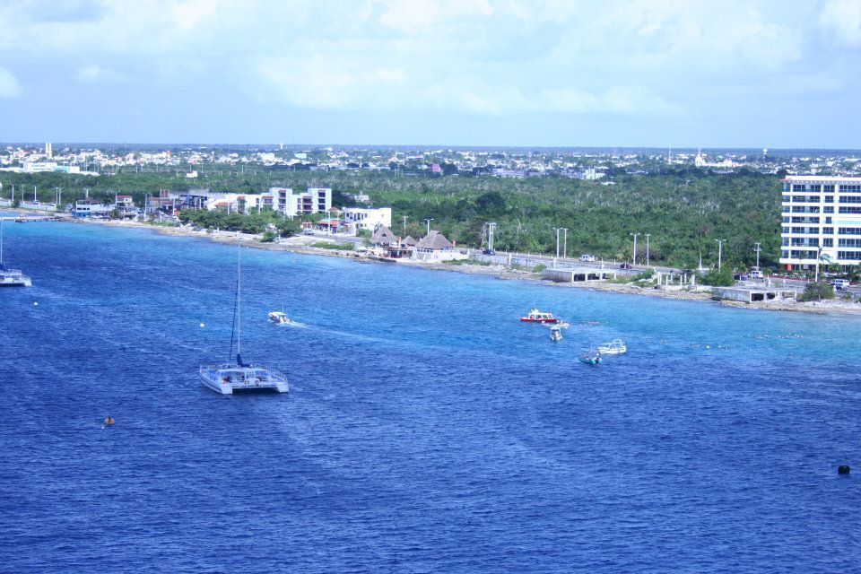 A coastal scene with blue water, boats, and a shoreline with buildings and vegetation under a cloudy sky.