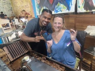 Man and woman smiling, holding cigars over a cigar-making machine, with art on the wall.