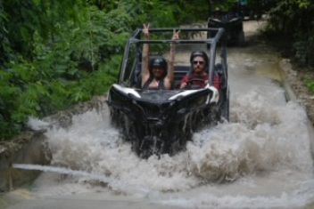Two people in an off-road vehicle splashing through water, surrounded by green foliage.