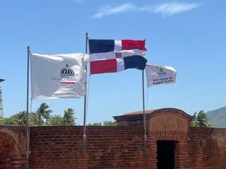 Three flags on a brick structure, including the Dominican Republic flag, flying under a blue sky.